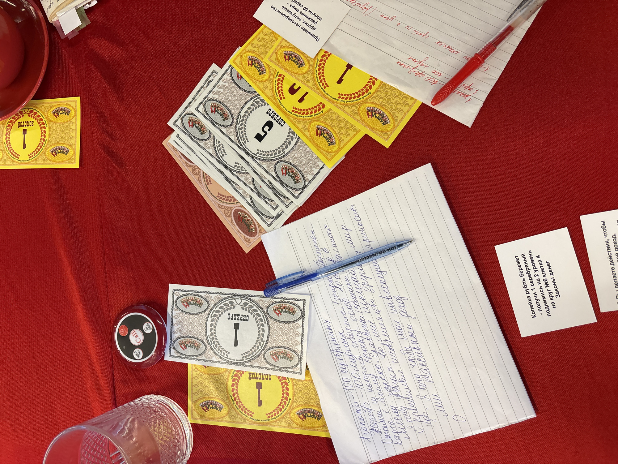 The image depicts a tabletop scene, likely set indoors, covered with a red cloth. Several items are scattered on the table, including:
* Play money resembling currency, some labeled "ЗолOTOE" (Gold) and "СЕРЕБРО" (Silver) with different denominations (1, 5, 10).
* A call button, potentially for service in a restaurant or similar setting.
* A clear drinking glass.
* Handwritten notes in Russian, containing information about game rules, tasks, and financial concepts like "passive income".
* Pens, both red and blue, used for writing the notes.
There is no direct indication of a location in Kuta Selatan, Indonesia, or any specific people present in the image. The scene appears to be a setup for a game or learning activity related to money, possibly involving role-playing or simulation.