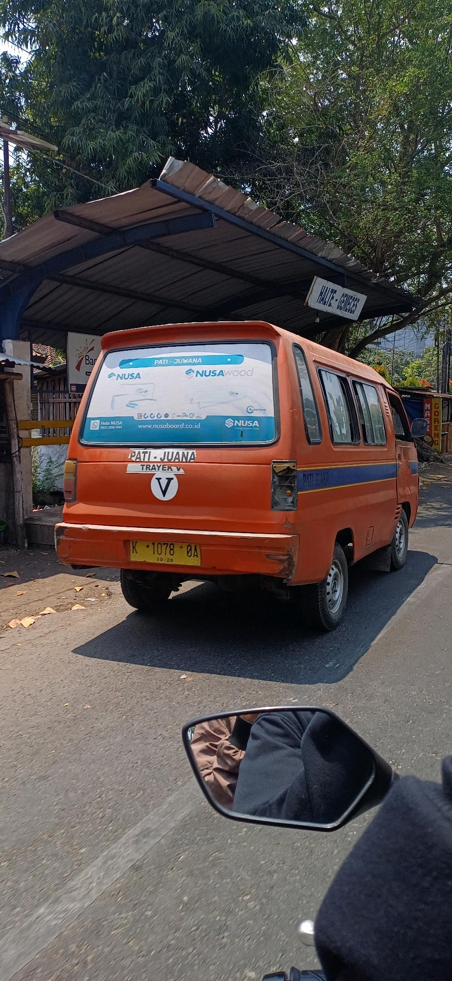 The image shows the rear of an orange mini-van with blue stripes, parked on a road in front of a shelter. The van has a sticker on the rear window advertising "NUSA" and "NUSWood" with a website address. Below the window, a plate reads "PATI - JUANA TRAYEK V" with a white circular emblem containing a black letter "V". A yellow license plate with black lettering "K. 1078. OA" and "10-26" is attached to the bumper. In the background, a sign reading "HALTE - GEMECES" is visible, indicating a bus stop. Trees and foliage are present around the scene, suggesting an outdoor, possibly rural or semi-urban environment. The lower portion of the image shows a reflection in a motorcycle's side mirror, revealing the rider's legs covered in dark clothing and a brown jacket. The road surface is asphalt. The overall lighting suggests it is daytime.