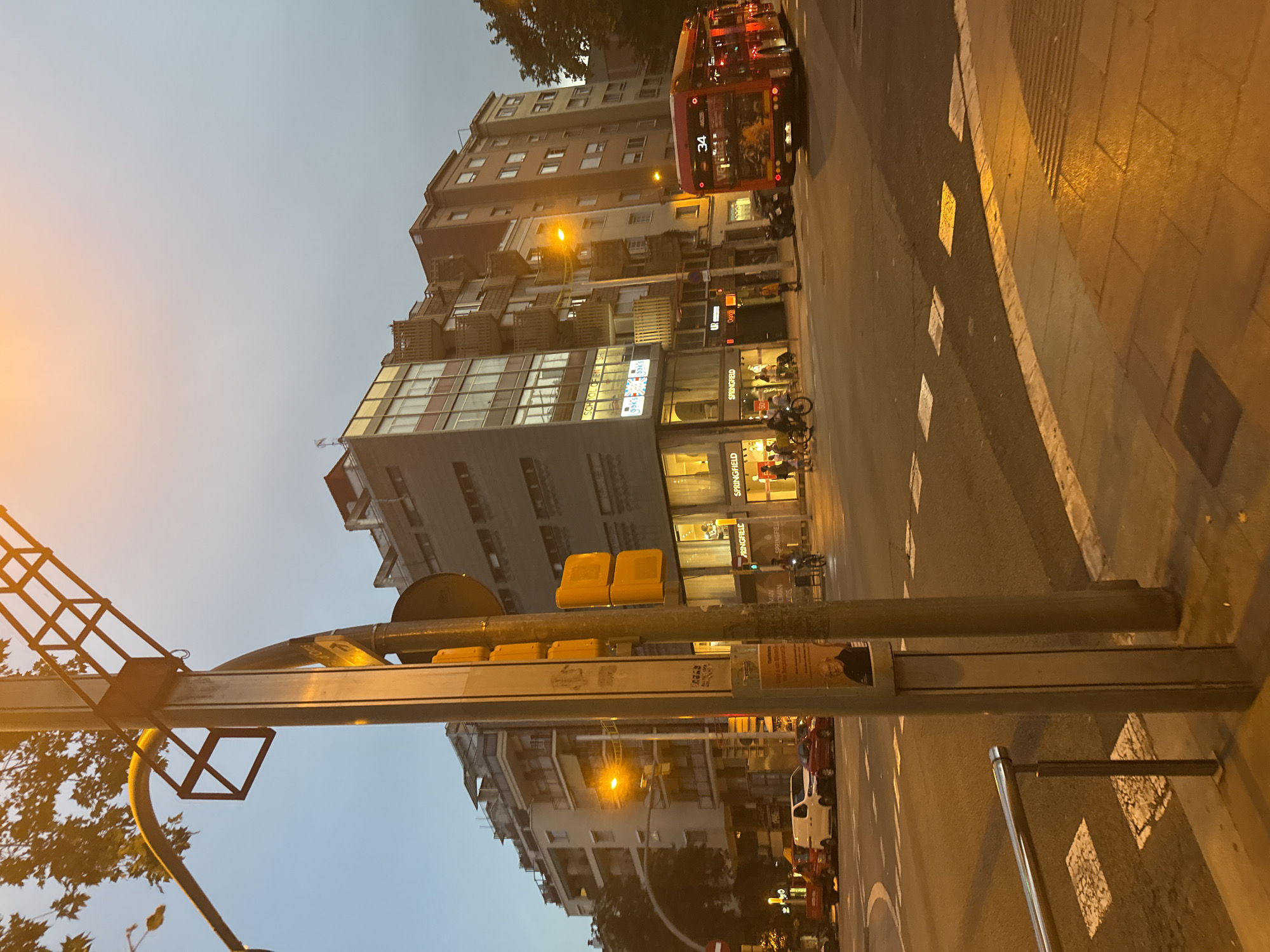 The image shows a street scene in Barcelona, Spain, at dusk. In the foreground, a metal railing is visible next to a crosswalk with white squares. A traffic light pole with yellow lights stands prominently to the right. Behind the pole, a red bus with the number 34 displayed on its front is traveling away from the camera.
The midground features a street with several cars, including a white van and a red car, parked on the left. Buildings line the street, with storefronts on the ground floors. Signs for "SPRINGFIELD" are visible. People are seen walking and cycling on the sidewalk.
In the background, multi-story buildings with balconies and windows dominate the skyline. Trees with foliage are also present. The sky is a pale blue with some orange hues, suggesting the setting sun. The overall impression is of a busy urban intersection during twilight.