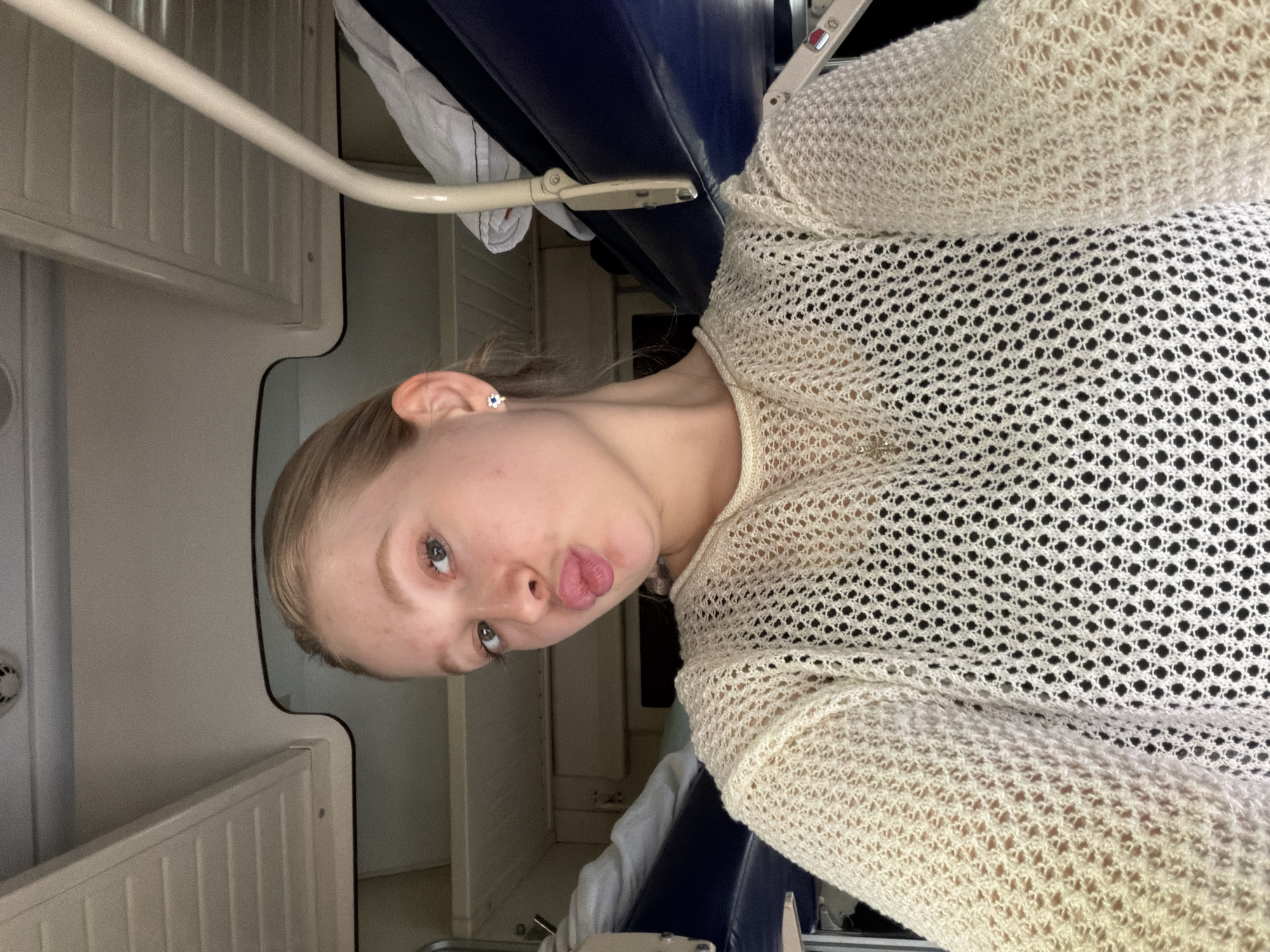 A young person with light skin and hair, wearing a white, crocheted-style sweater, is inside what appears to be a train compartment. The walls are pale with paneling, and a luggage rack is visible overhead. The person is sticking out their tongue. The image may have been taken in Зелёный Дол, Russia, as specified by the prompt.