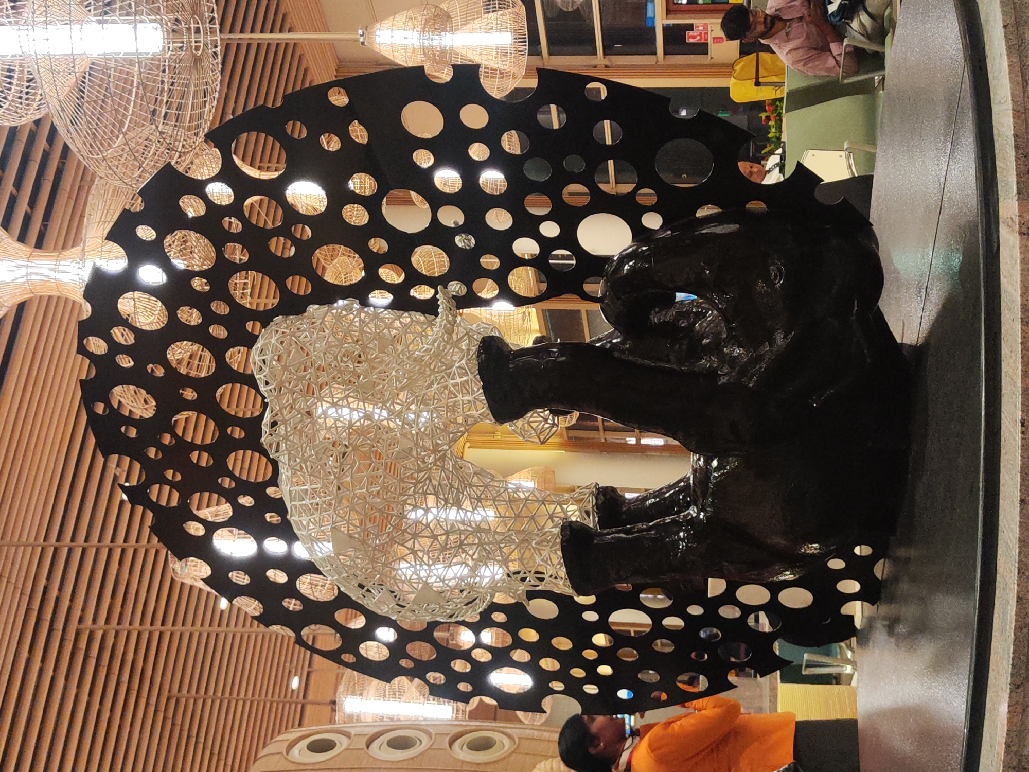 Captured indoors, the photograph features a distinctive artwork in Gangamuthanahalli, India. The artwork involves a large black structure with numerous circular cutouts, positioned behind a white, lattice-like spherical form. Juxtaposed is a dark sculpture resembling an elephant, resting on a dark, reflective surface near a grey, textured wall. The backdrop showcases a wooden ceiling with woven light fixtures. People are visible in the scene: one individual dressed in orange clothing and another seated and facing away from the camera. Additional seating is observable in the periphery.