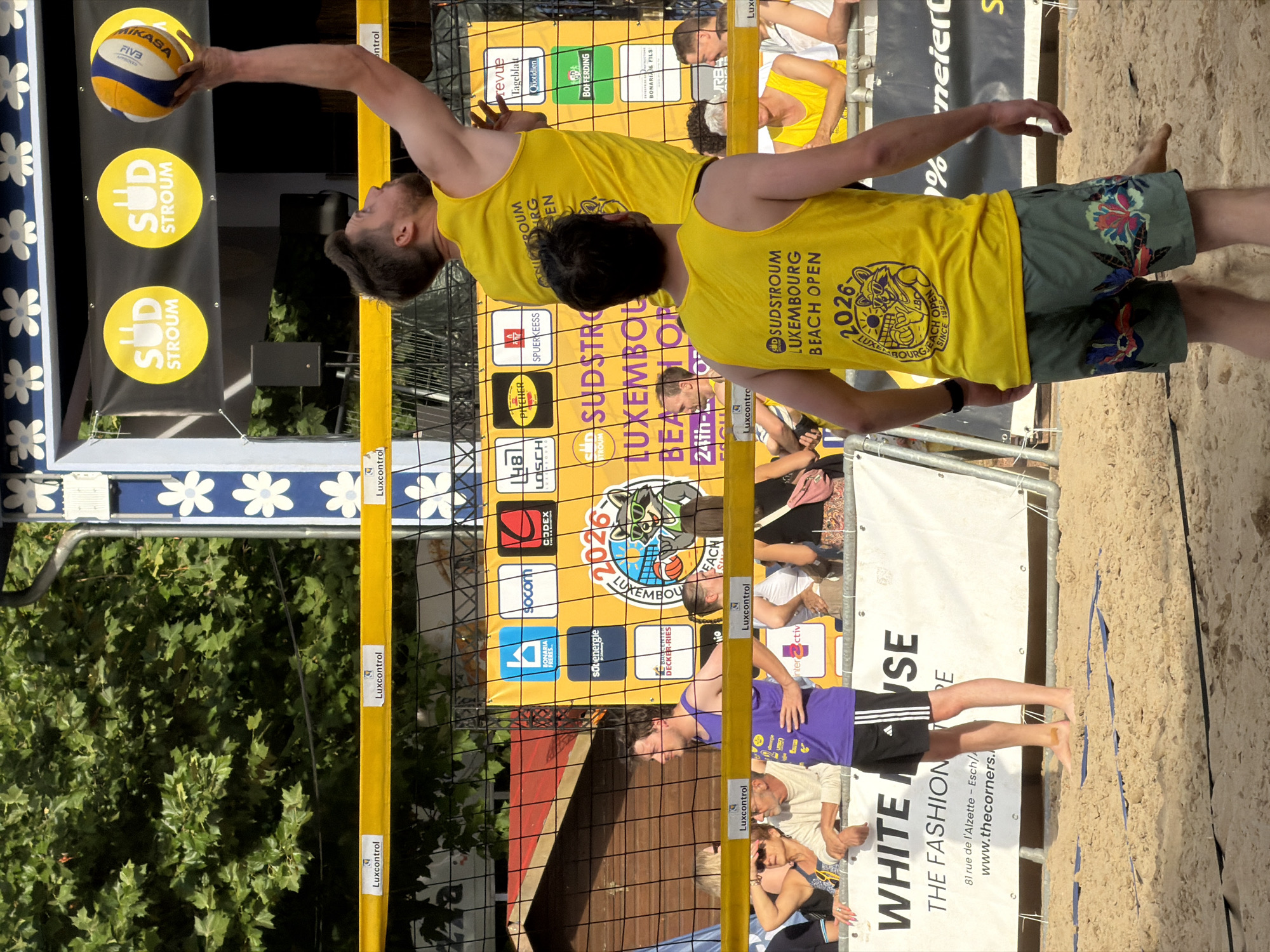 The image depicts a beach volleyball match at the "Sudstroum Luxembourg Beach Open 2026" event, likely held in Esch-sur-Alzette, Luxembourg, given the prominent sponsor "White House, The Fashion House" and the address "81 rue de l'Alzette - Esch". In the foreground, a male volleyball player wearing a yellow tank top and floral patterned shorts is positioned on the sand, facing away from the camera. He appears to be in a relaxed stance, possibly waiting for play to resume. Behind him, another male player in a yellow tank top with "Sudstroum Luxembourg Beach Open" text is jumping with a volleyball in his raised hand, suggesting an action shot of a serve or hit. A net separates the players. In the background, spectators are visible behind a barrier. The environment is a sandy beach volleyball court under a bright sky with trees and structures, including advertising banners, visible behind the court.