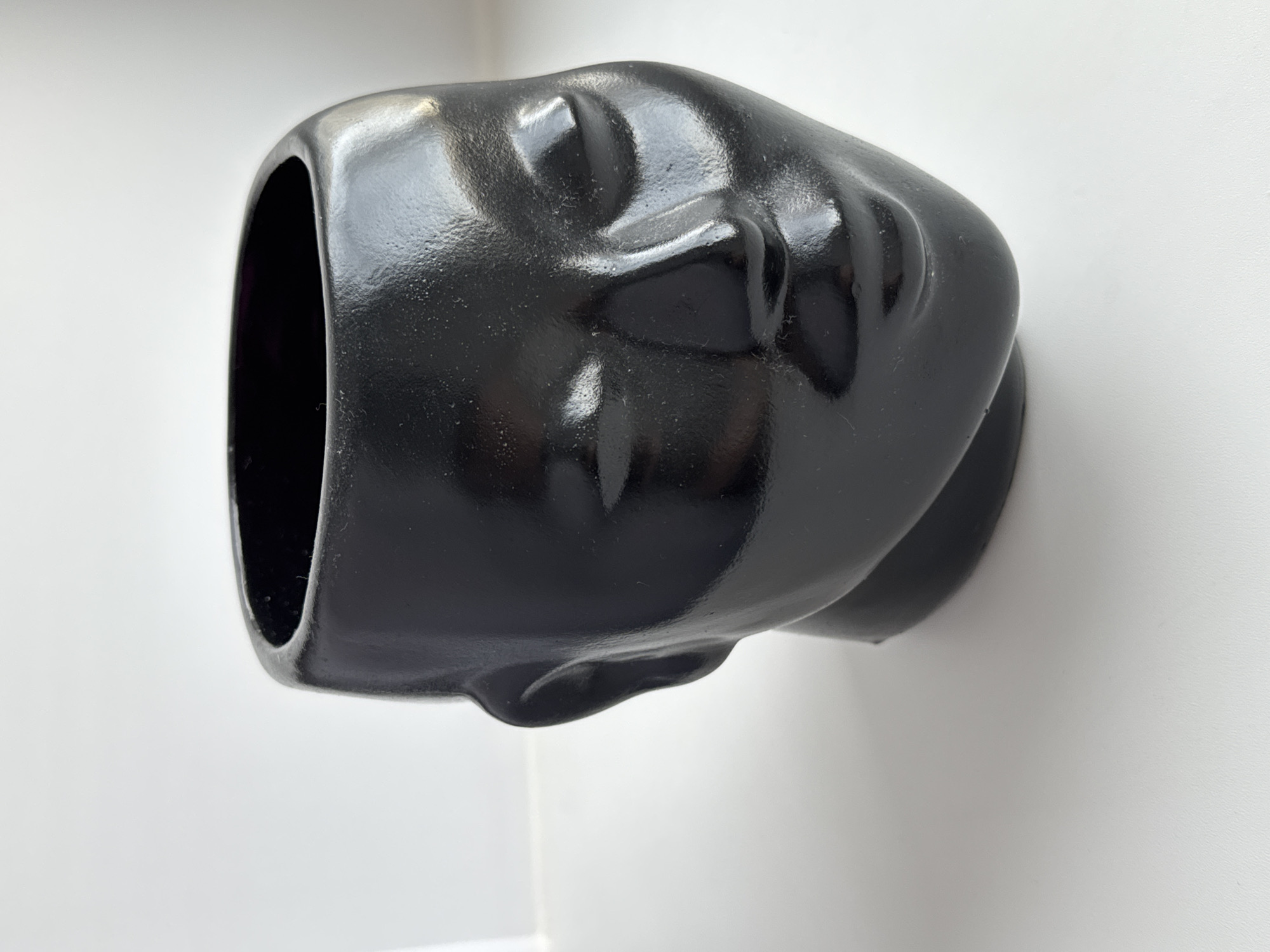 A black ceramic head with an open top, reminiscent of a planter or vase, is positioned against a plain white wall. The head is sculpted with subtle facial features. The location and environment are otherwise indeterminate.
