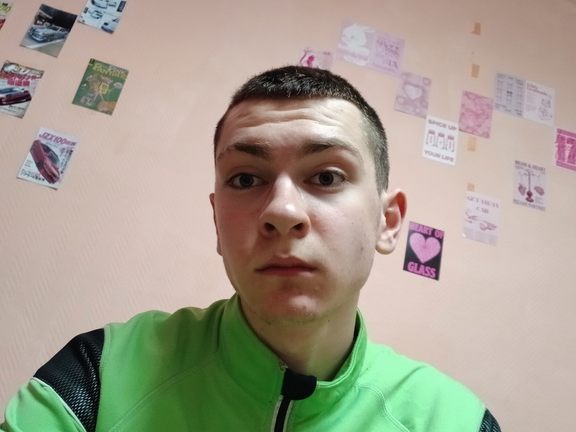 A close-up selfie of a young Caucasian male with a buzzcut haircut. He has light skin, dark hair, and a neutral expression. He is wearing a lime green and black athletic jacket.
The background is a pale peach-colored wall adorned with various small posters and pictures, including images of cars, a Pink Panther cartoon, and motivational phrases. The location is not readily discernible as Chaikovsky, Russia from the image itself.