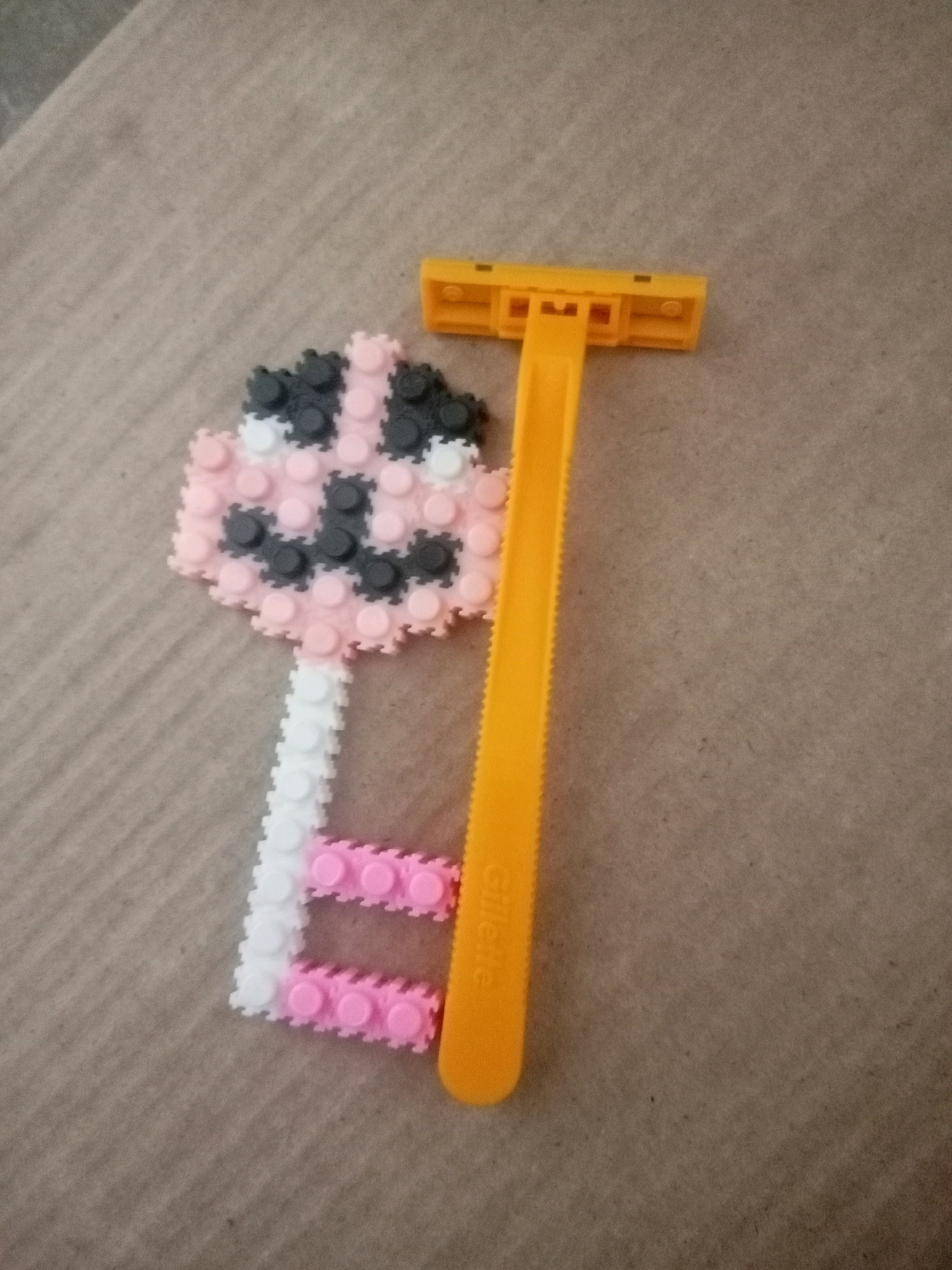 A close-up, high-angle shot shows a yellow plastic razor positioned vertically to the right of a key-shaped object constructed from colorful small interlocking plastic bricks. The key-shaped object has a round head featuring pink, white, and black bricks arranged in a pattern that resembles a face. The stem of the key is made of white bricks, with two horizontal pink brick segments forming the end of the stem. The razor has a textured grip and the brand name "Gillette" is visible in white text on the orange handle. The objects are placed on a textured, neutral-colored surface, possibly a mat or fabric. This image was taken in Batam, Indonesia.