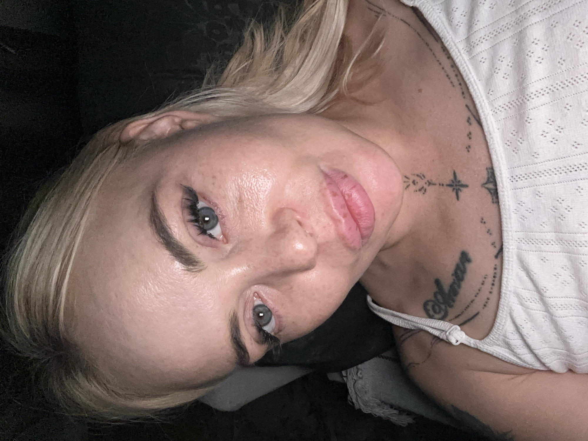 A female individual is depicted in a close-up, head-and-shoulders portrait, positioned against a dark, indistinct background in an unknown indoor location. The subject has blonde hair, light blue-grey eyes looking directly at the viewer, and fair skin with visible texture and scattered freckles. Her lips are full and appear slightly glossy, with several small reddish marks on the lower lip. She is wearing a white, textured camisole with thin straps. Visible tattoos include cursive script on her right upper chest/collarbone area and a vertical, geometric design on her lower neck and upper chest, incorporating a star-like element and linear patterns. A light-colored, textured fabric is partially visible on the left side of the frame in the background. The lighting appears to be artificial, illuminating the subject's face.