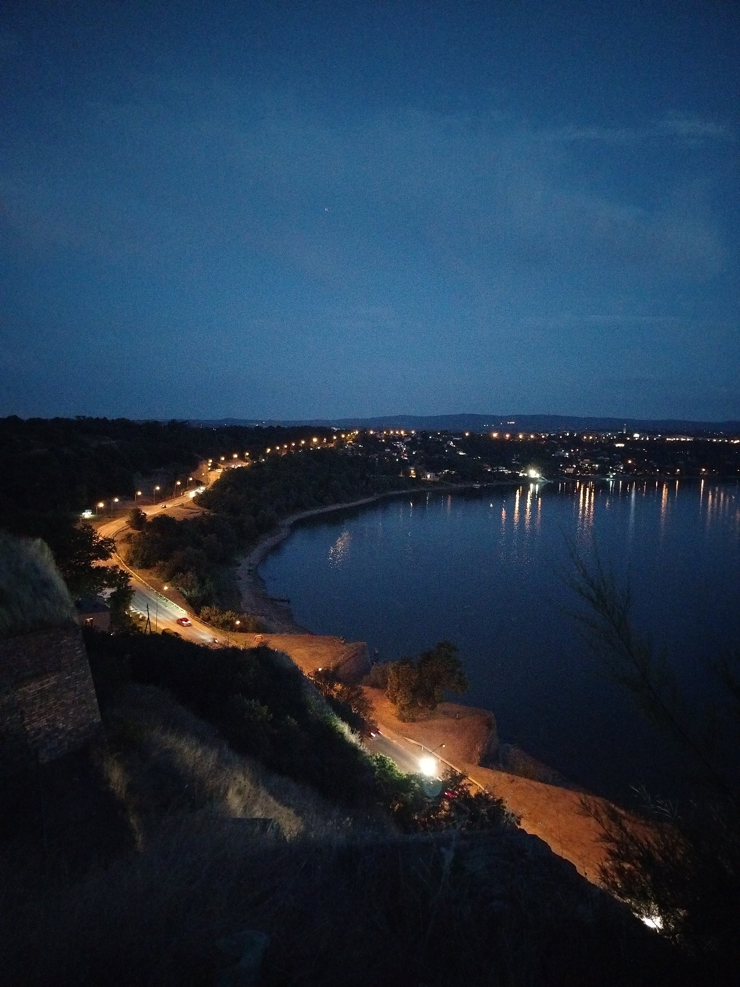 This image captures a twilight scene from the Petrovaradin Fortress in Petrovaradin, Serbia. The viewpoint is elevated, looking down and across a body of water, likely the Danube River. A winding road is illuminated by streetlights, with a few cars visible in motion. On the opposite bank, a city skyline is visible, dotted with lights, suggesting it is the city of Novi Sad. The water reflects the city lights and the streetlights from the road. The foreground shows a dark, textured area, likely part of the fortress's ramparts or landscaping, with some sparse vegetation. The sky is a deep blue, indicative of the fading light after sunset.