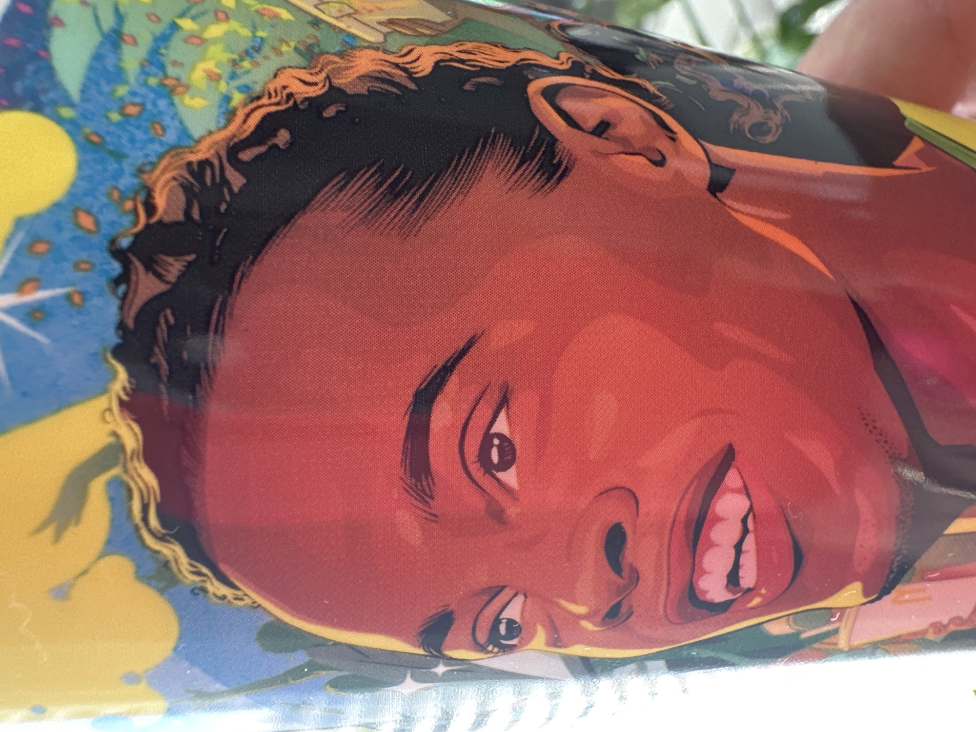 The image is a close-up shot of a cup featuring an illustration of a young Black person's face. The illustration is in a stylized, comic book-like art style with bold colors and sharp lines. The person's skin is rendered in vibrant reds and oranges, with dark eyebrows and short, curly black hair. Their eyes are looking slightly to the left, and they have an open-mouthed smile, revealing white teeth. The background of the cup is a colorful abstract design with elements of blue sky, yellow shapes, and hints of green foliage. The cup is held by a hand, with some of the background environment, possibly foliage, visible out of focus. The image is taken from a slightly upward angle.