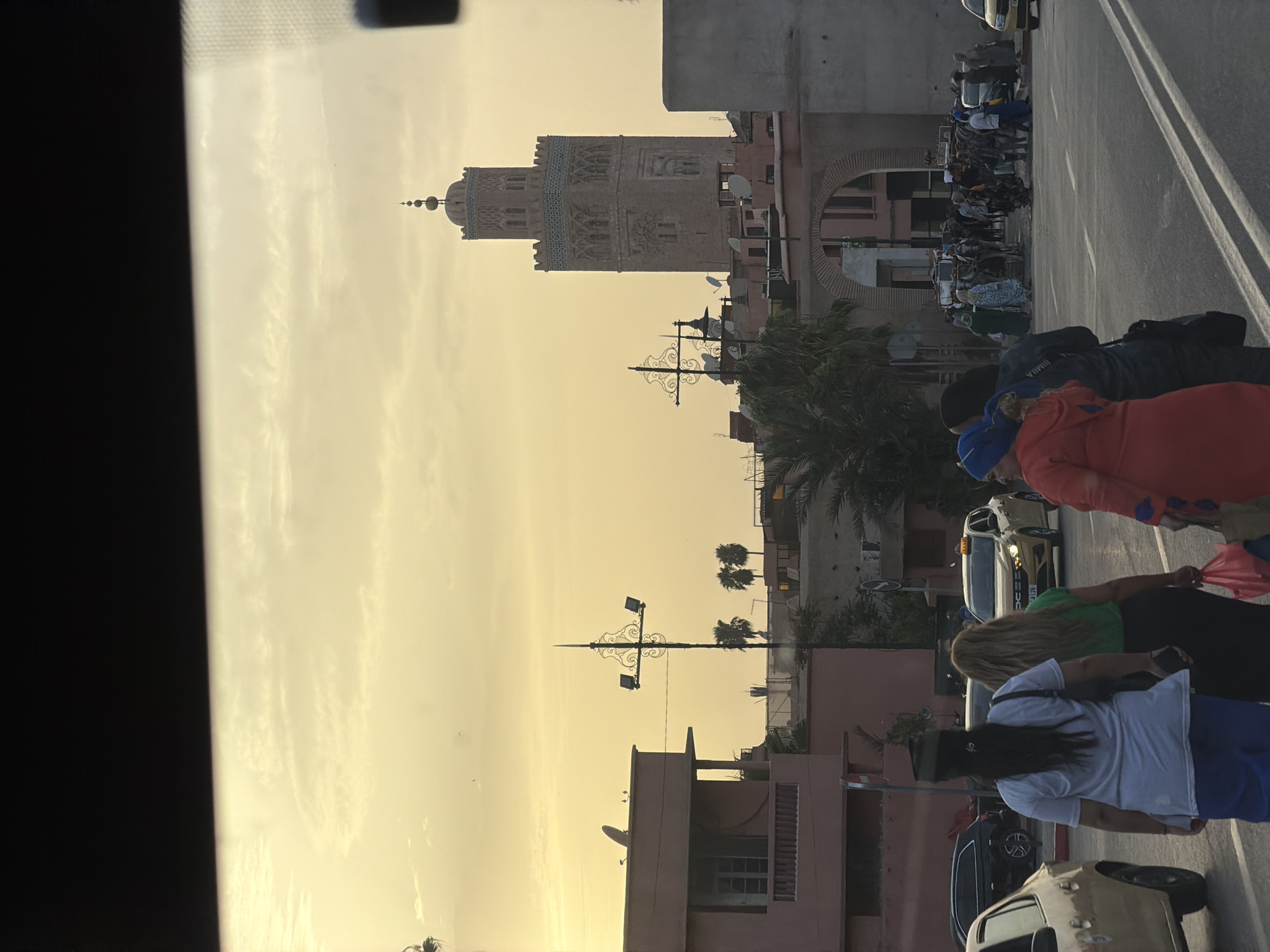 This is a photograph of a street scene in Marrakesh, Morocco, with the Koutoubia Mosque minaret prominently visible in the background under a hazy, yellow sky. Several people are walking on the street and sidewalk, some carrying bags. A yellow taxi is driving on the road. Buildings with pink facades and balconies line the street, and palm trees are visible. An arched gateway is also present in the scene. The time of day appears to be late afternoon or early evening, indicated by the warm, soft light.