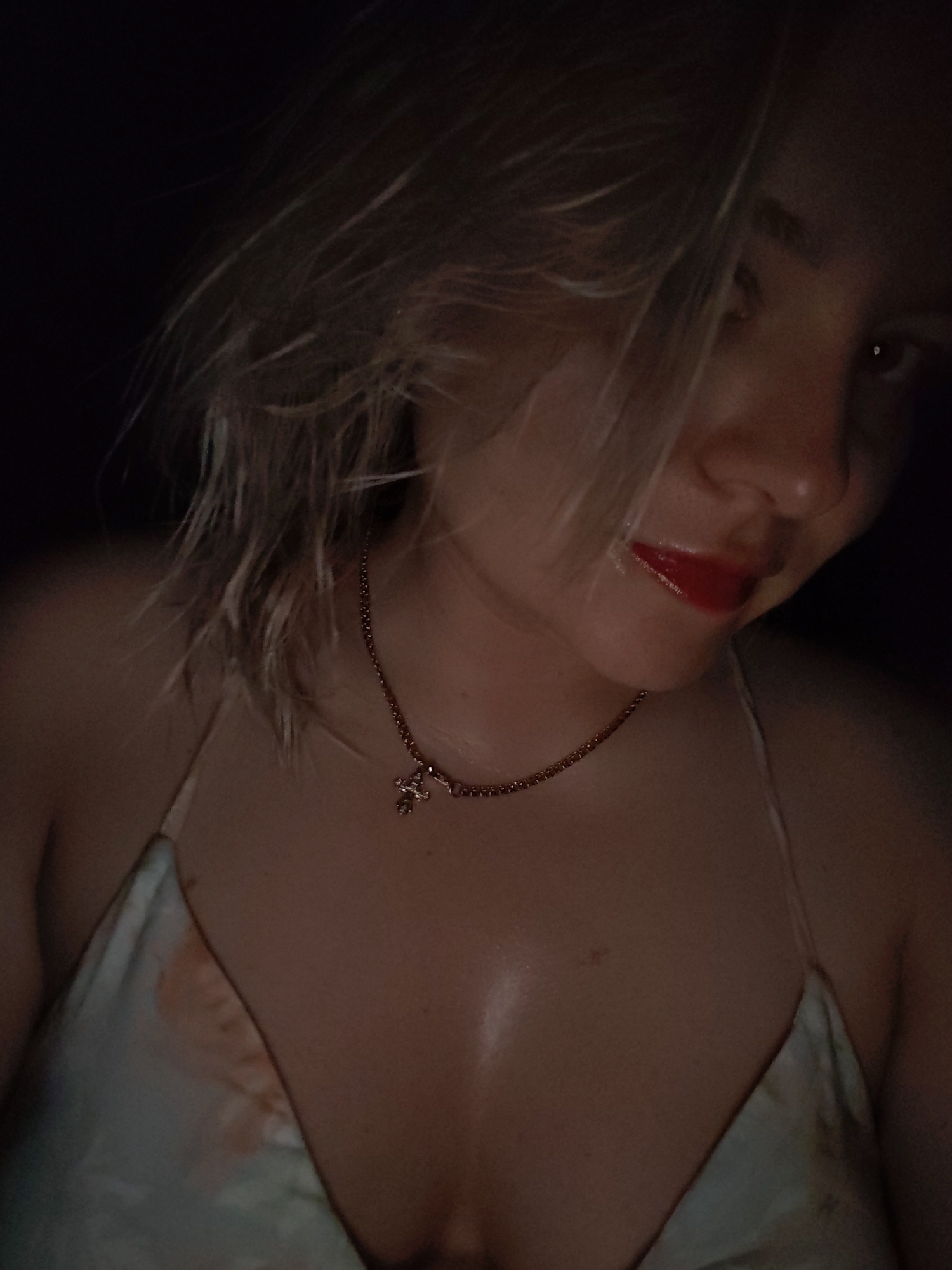 A close-up shot shows a woman with blonde, damp hair, wearing red lipstick and a light-colored bikini top with a delicate gold necklace featuring a cross pendant. The background is dark, obscuring details. The image captures the woman from her shoulders up, with a slight downward tilt of her head. The lighting is dim, casting shadows and highlighting her features. There's no information to ascertain the location of the woman or any information about the city of Izhevsk, Russia.