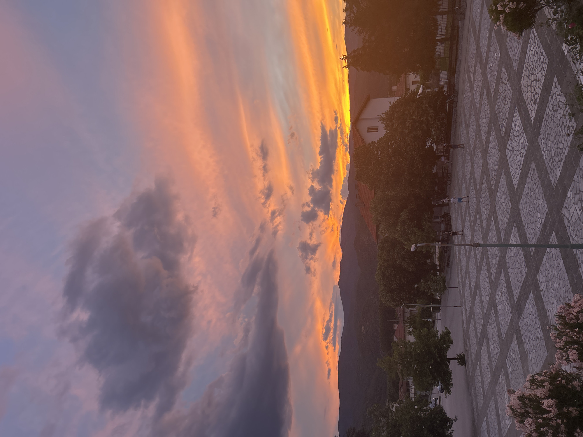 The media file depicts a sunset over a town square. The sky is ablaze with streaks of orange, pink, and yellow, with darker clouds tinged with purple and grey. Mountains form a silhouette against the vibrant sky on the right side of the frame. In the foreground, a patterned stone plaza is visible, with a street lamp and trees lining the edge. Several people are scattered throughout the plaza, some walking and others seemingly standing or playing. A few buildings and more trees are visible behind the plaza, suggesting an urban or semi-urban setting. The warm glow of the sunset suggests it is late evening. The overall mood appears peaceful and serene, with the natural beauty of the sunset contrasting with the man-made environment of the plaza. There is no visible text in the image.