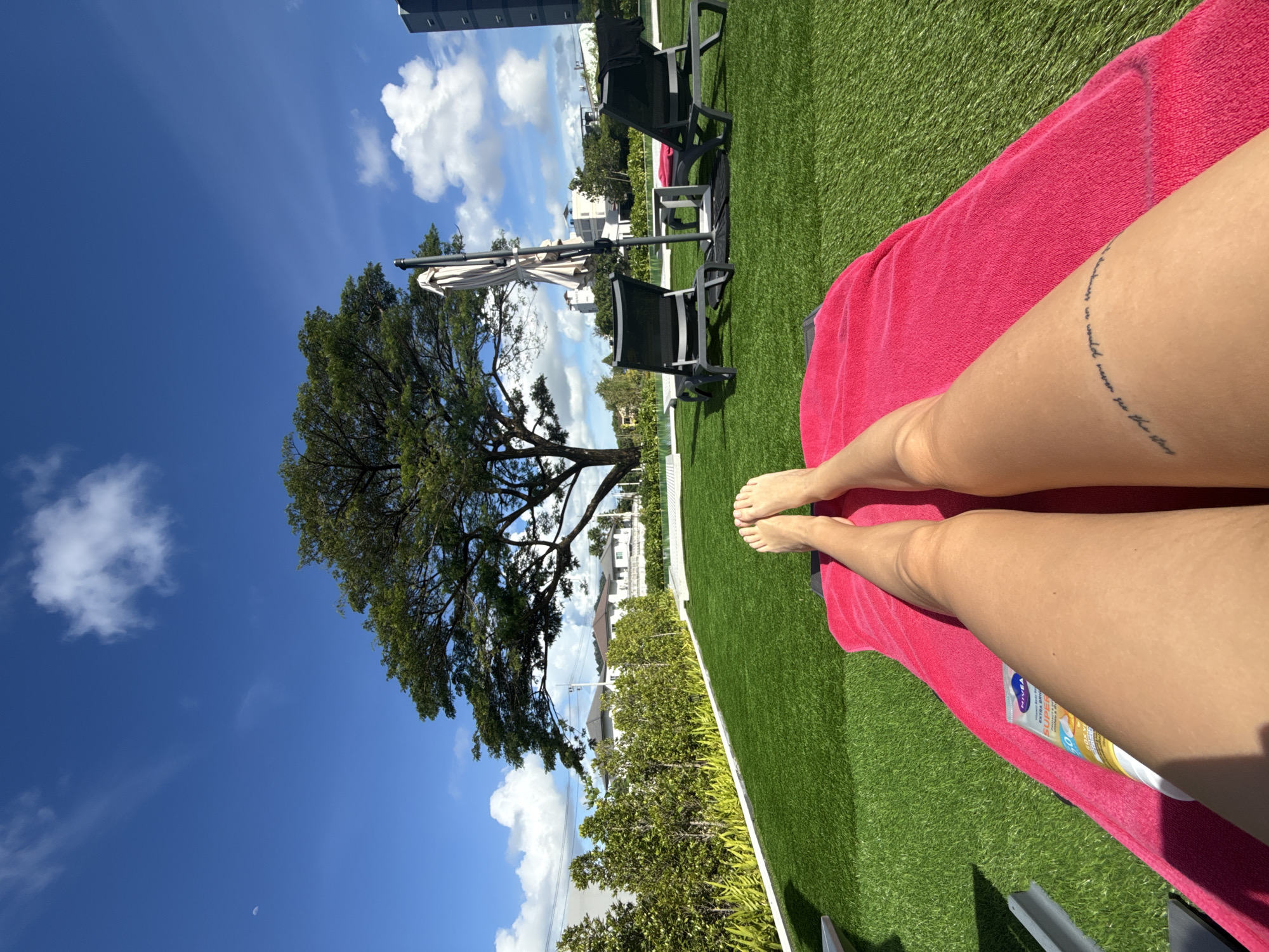 A first-person perspective shot shows a pair of tanned legs resting on a bright pink towel spread out on an artificial green lawn. The legs have a faint curved tattoo on the left thigh. In the foreground, a tube of Nivea sunscreen is visible. The background features two empty black lounge chairs next to a large patio umbrella. Beyond the chairs, lush green bushes line a white border. In the distance, several white houses with tiled roofs are visible, along with a large tree with a full canopy. A tall, dark building stands to the right of the frame. The sky is a vibrant blue with scattered white clouds and a sliver of the moon visible in the upper left corner. The scene is likely a resort or hotel pool area in Ratsada, Thailand, given the sunny weather and tropical vegetation.
