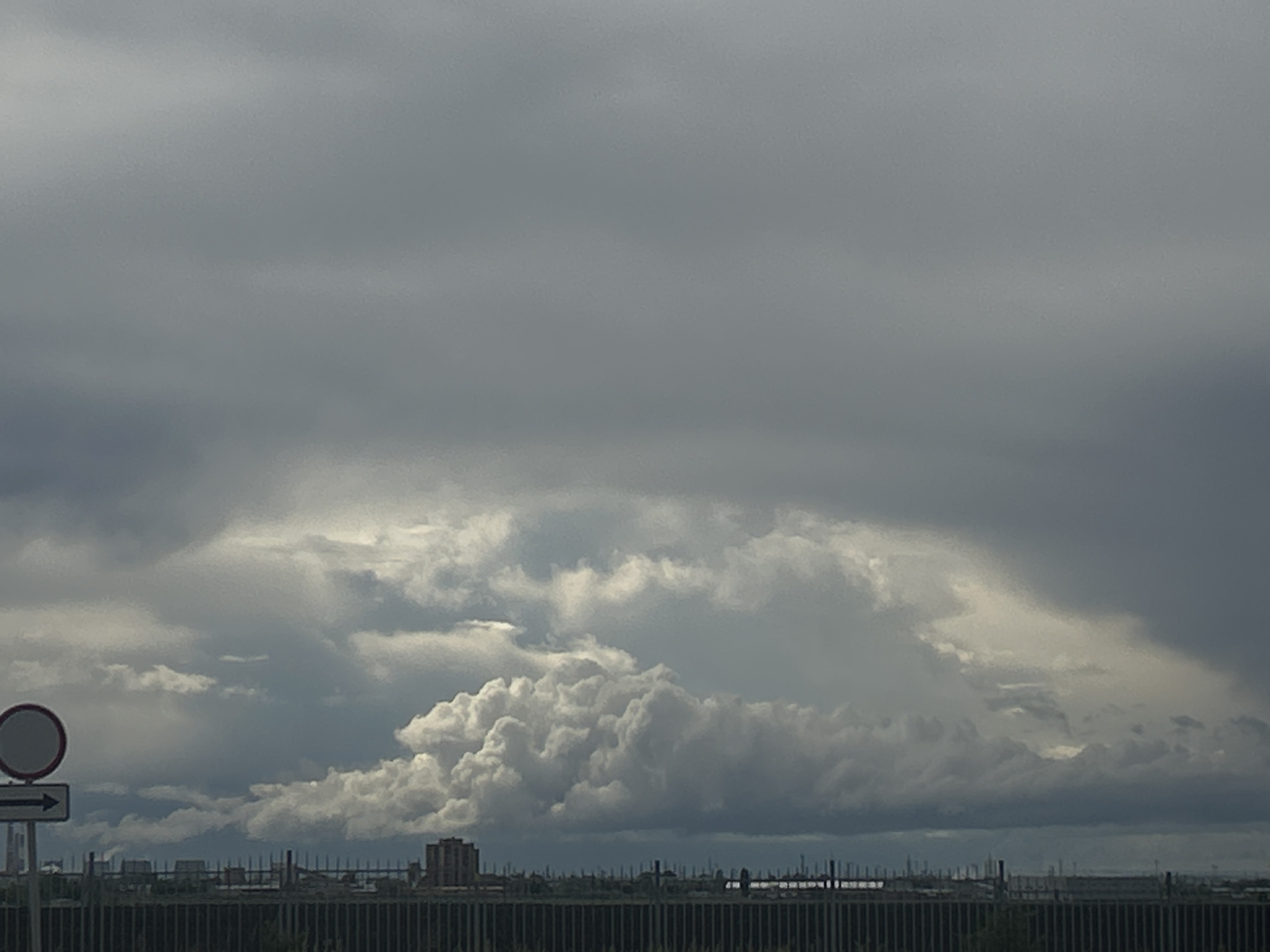 The image shows an overcast sky with a large formation of cumulonimbus clouds. In the foreground, there is a road sign with a white circle and a horizontal arrow pointing to the right. Behind the sign, a fence runs across the lower part of the frame, obscuring the horizon. Beyond the fence, there is a cityscape with buildings and industrial structures. Given the location in Тимофеевка, Russia, the industrial features suggest a possible urban setting. The absence of people and the focus on the environmental and man-made elements create a stark, descriptive portrayal of the scene.