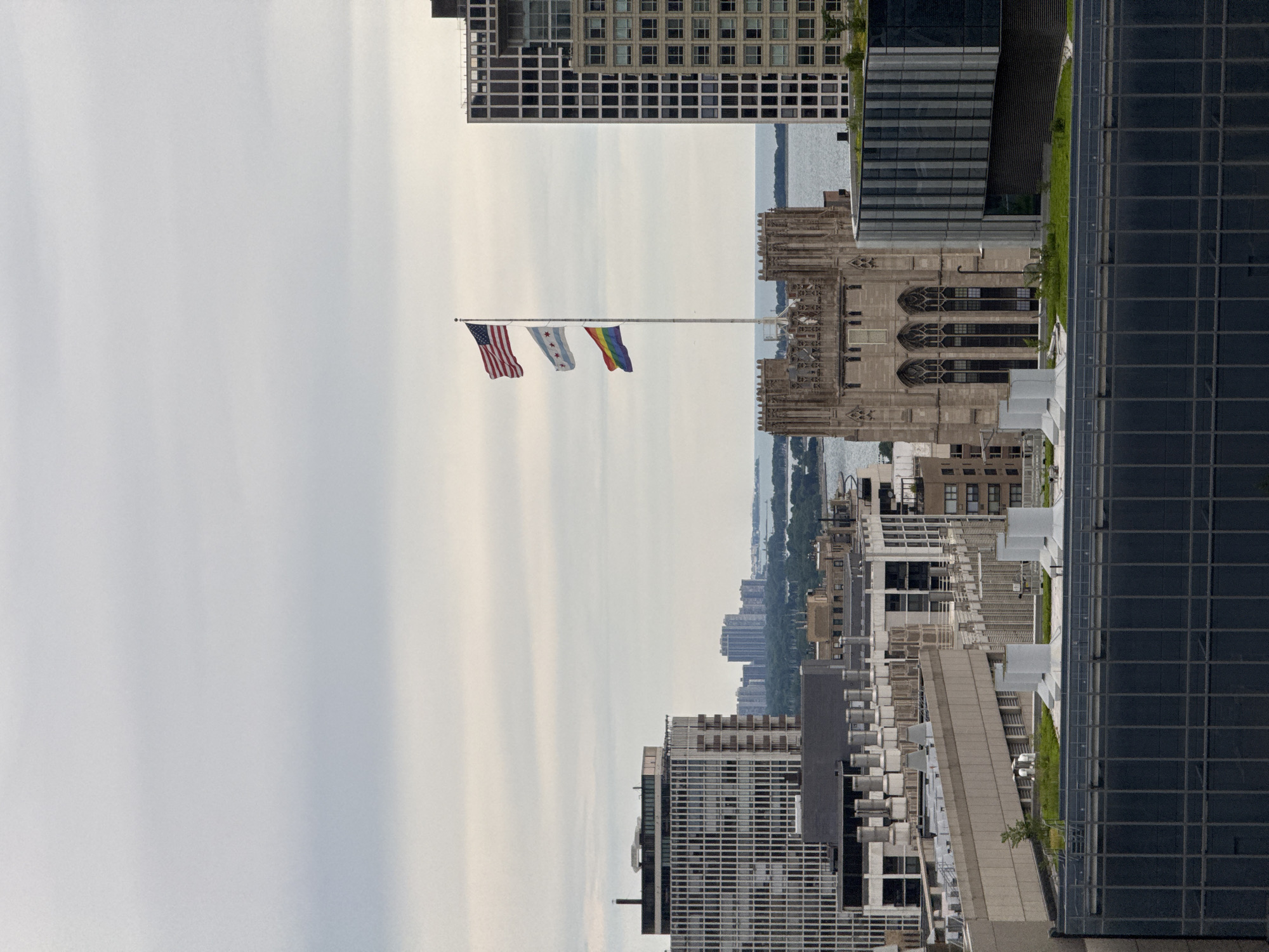 This is a medium-angle, vertically oriented, aerial photograph taken from a high vantage point, likely a skyscraper in Chicago. The scene captures a cityscape under a cloudy sky, with an American flag and two other flags flying on a flagpole in the foreground. The flags are positioned to the left of a Gothic-style building with intricate stone carvings and arched windows.
Beyond the immediate buildings, a body of water, likely Lake Michigan, is visible, stretching towards a hazy horizon lined with distant city buildings. The architecture of the city is a mix of modern glass towers and older, more ornate structures. Some buildings feature green rooftops, suggesting the presence of rooftop gardens.
The lighting suggests it is daytime, possibly overcast or nearing dusk, due to the diffused light and muted colors. No people are clearly discernible in the frame, but the flags indicate a civic or possibly a celebratory context. The orientation of the image is rotated 90 degrees clockwise from the standard vertical orientation, so the flags are flying horizontally across the frame. No text is visible in the image.