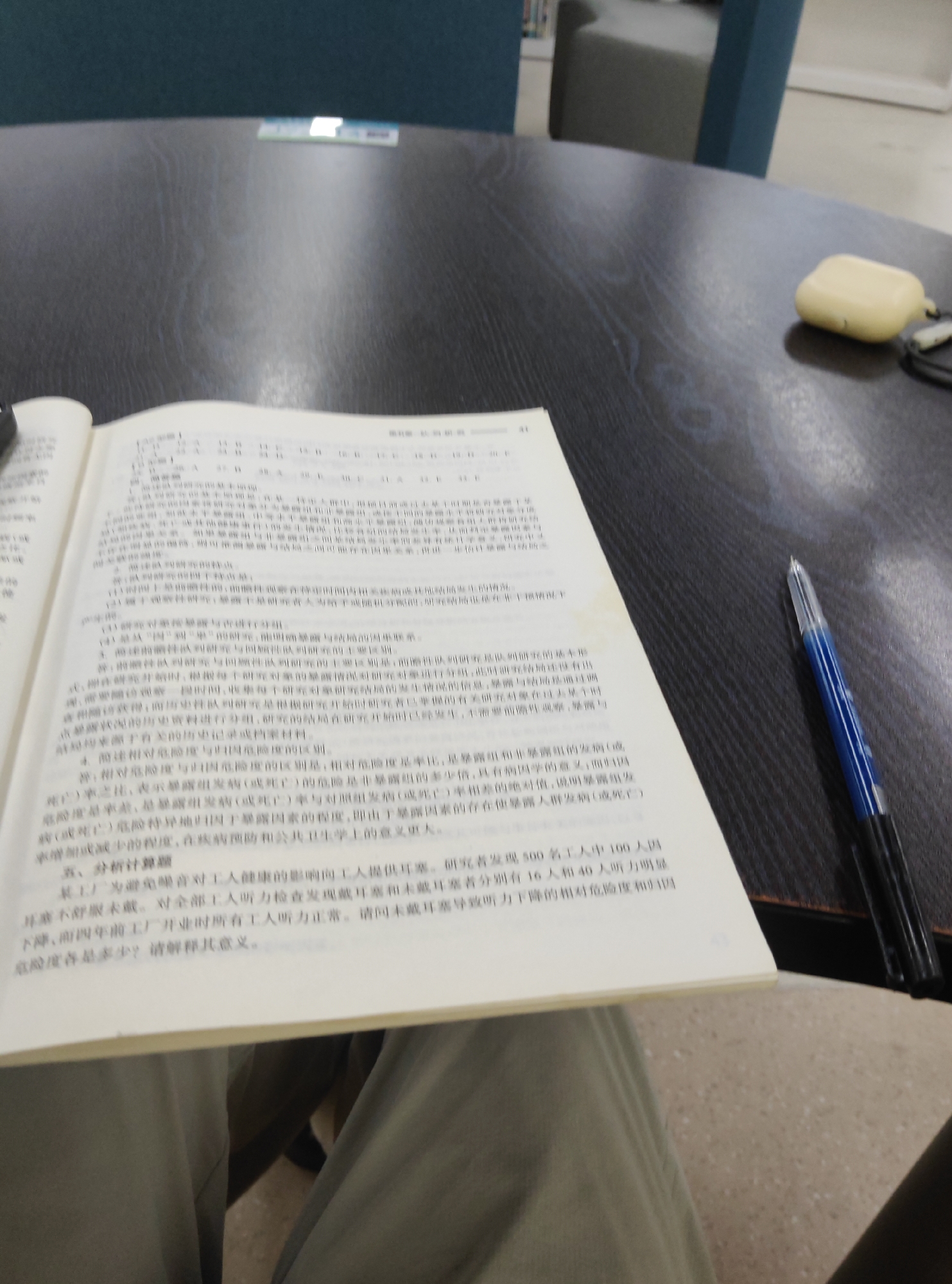 The image shows an open book with Chinese text on a dark, textured surface. A blue and black pen rests on the right side of the book. A cream-colored case for earbuds is visible to the right of the book, on the same surface. The background is blurred, but appears to be an indoor setting with furniture, possibly a library or study area. There is no visible indication of the location being the Chongqing High-tech Zone, China, nor are there any people or specific actions depicted. The scene is static and focuses on the book and writing implement.