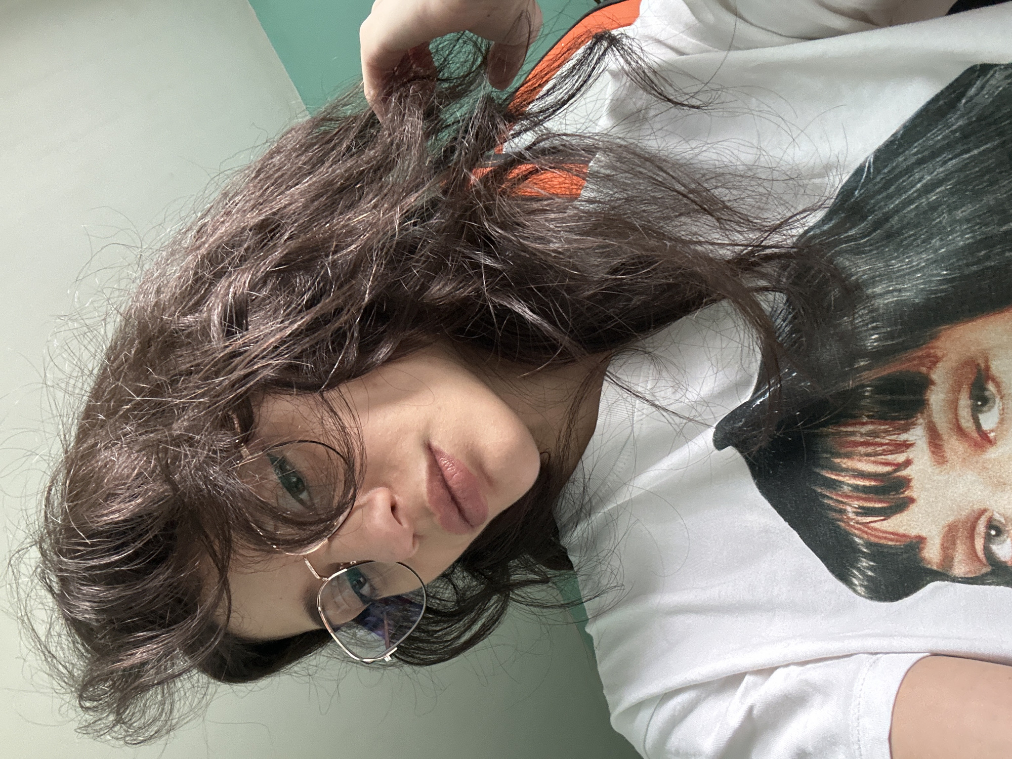 A young woman with long, dark, messy hair and glasses is taking a selfie in a room. She is wearing a white t-shirt with a picture of a woman's face on it. The background is a light-colored wall with a teal accent wall on the right. The image does not contain any information about the city of Sofia, Bulgaria.