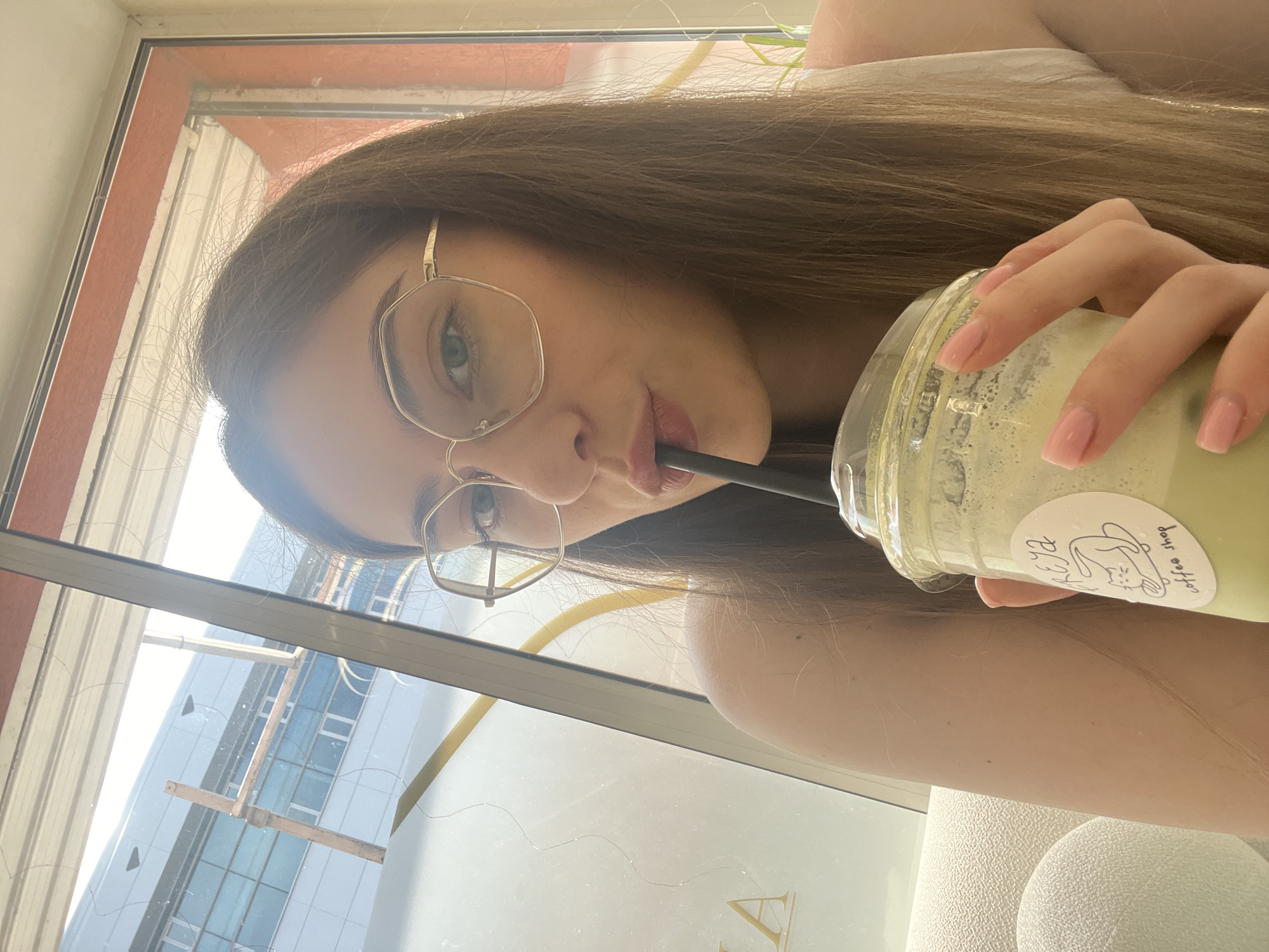 A close-up, high-angle portrait shot shows a person, likely female, holding and drinking from a beverage cup while seated indoors by a large window. The image is taken from a slightly upward perspective, with the person looking directly at the camera.
The subject has long, straight brown hair that cascades over their shoulder and is parted in the middle. They wear large, hexagonal gold-rimmed glasses, which frame their light-colored (possibly hazel or green) eyes. Their lips are slightly parted around a black straw, suggesting they are actively drinking. A hint of light pink lip color is visible. One arm is visible, showing clear, light pink manicured fingernails grasping a transparent plastic cup.
The cup contains a light green, frothy beverage, likely a matcha latte or similar drink. A white oval sticker is adhered to the cup, featuring a minimalist drawing that resembles a cat or bunny head, along with handwritten text that appears to read "Elysa" or "Elysia" and "coffee shop".
The setting appears to be a cafe or coffee shop during the daytime, indicated by the bright light streaming through the large window behind the person. Through the window, modern buildings with reflective surfaces and some scaffolding or structural elements are visible. Inside, a glimpse of a light-colored, textured chair or seating is seen on the right, and a gold-colored decorative element is visible through the lower part of the window. The overall mood is casual and relaxed.