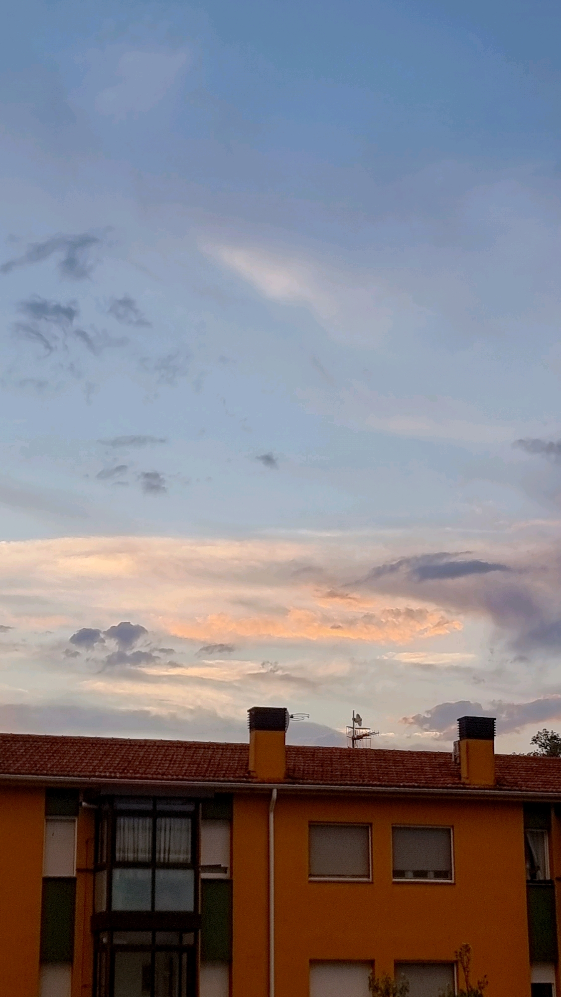 The image depicts a section of a building in Sant Joan Les Fonts, Spain, with a sky full of clouds during sunset. The building has a prominent orange facade with a tiled red roof. Two chimneys, each with black tops, are visible on the roof. There are several windows on the building, some with shutters. A glass-enclosed balcony area is also present. The sky above transitions from a pale blue at the top to softer hues of pink and orange near the horizon, indicating the sunset. Wispy clouds are scattered across the sky, with darker, more defined clouds clustered lower down. No people or animals are visible in the frame. The environment suggests a residential or commercial building in a town or city.