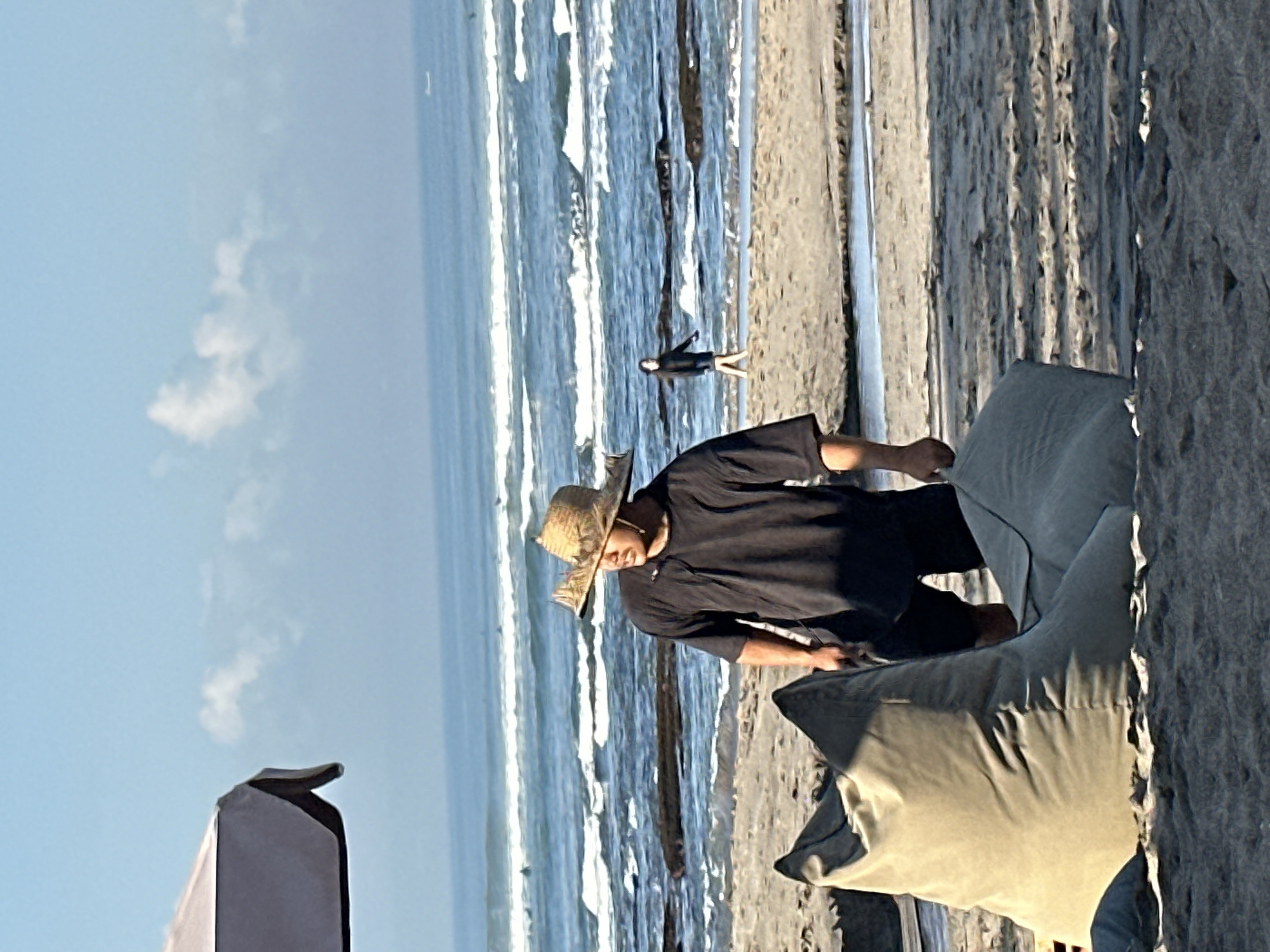 A man wearing a straw hat and a black t-shirt stands on a sandy beach in North Kuta, Indonesia, facing the ocean. He is partially obscured by a large, olive-green beanbag chair in the foreground. Another beanbag chair is visible to the right. In the background, the ocean has moderate waves, and several surfers are in the water. Further in the distance, another person is walking on the beach near the water's edge. A large umbrella is visible on the left side of the frame, and the sky is clear with a few scattered clouds.