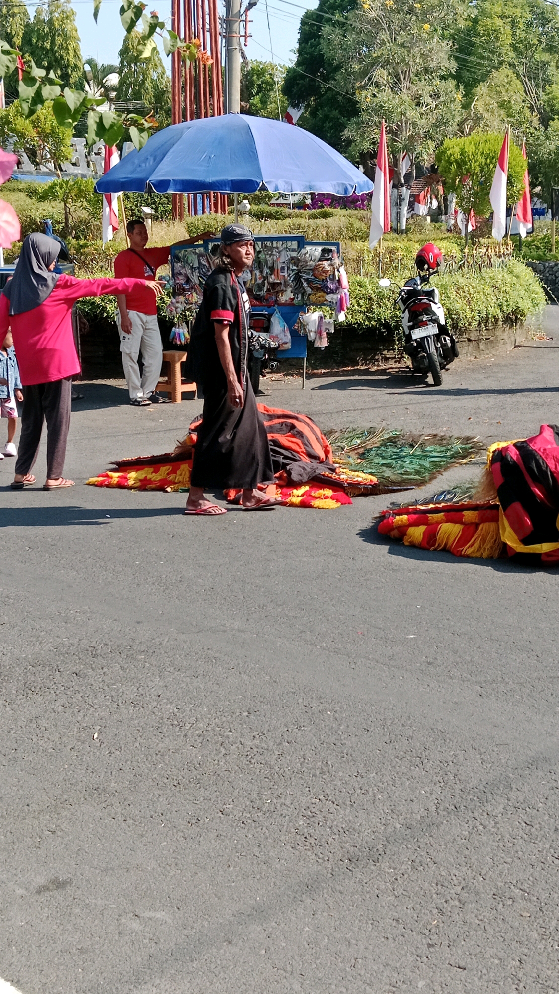 In Magetan, Indonesia, a street scene unfolds under a bright, sunny sky. A woman in a pink headscarf and long-sleeved shirt gestures to the left, her dark pants and sandals indicating she is standing on asphalt. Beside her, a man in a red t-shirt and white pants stands near a blue umbrella and a vendor's cart laden with goods. To the right, a man in a black robe and headscarf walks forward, his sandals on the asphalt. In front of him, colorful, ornate costumes, possibly for a performance, lie on the ground, one resembling a lion's head. A red-helmeted motorcycle is parked on the right side of the scene, near a hedge and Indonesian flags adorning poles. The background shows trees and buildings.