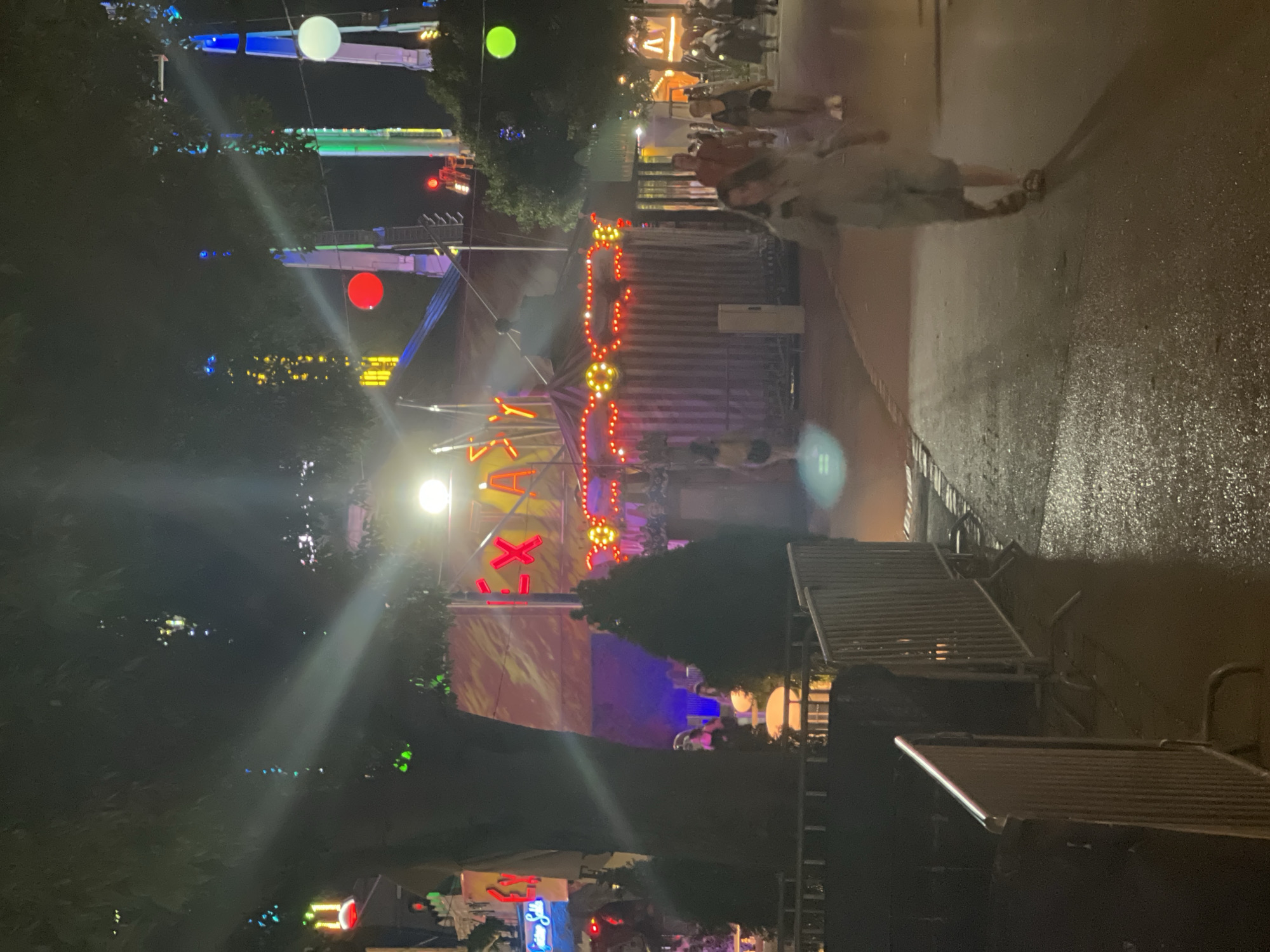 The image depicts a nighttime scene at a carnival or fairground, likely in Vienna, Austria, given the context provided. Several amusement rides are visible, including tall, illuminated structures with colored lights, and what appears to be a carousel or a similar attraction with red and yellow lights. The ground is wet, suggesting recent rain or maintenance, reflecting the bright lights of the attractions. Numerous people are walking along a paved pathway, some blurred due to motion. One individual in the foreground is walking away from the camera, wearing light-colored clothing. In the background, a large tent-like structure with neon red lettering that reads "EXA.." is prominent. The atmosphere is festive, with a variety of colored lights creating a vibrant nighttime ambiance.