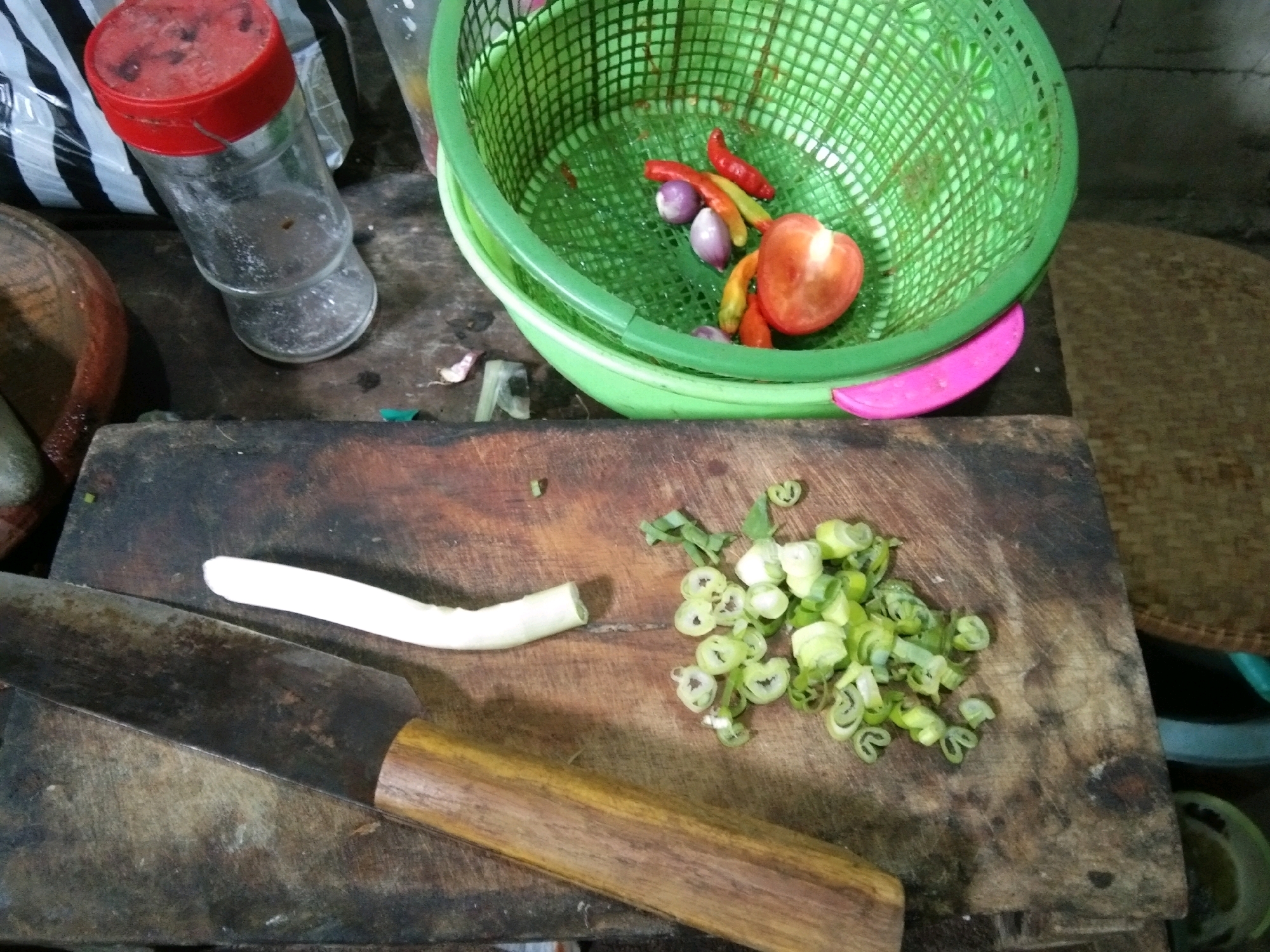 A close-up, overhead shot captures a culinary scene on a wooden cutting board. To the left, a large knife with a worn wooden handle rests horizontally across the board, its blade extending out of frame. Next to the knife, a white stalk, likely a leek or large spring onion, is cut in half, with the larger portion still on the board. To the right, a pile of chopped green onions, sliced into small rounds with visible white stems and green tops, is scattered across the board. In the background, a green plastic colander holds red chilies, two small purple onions, and a halved tomato, suggesting ingredients for a meal. A glass jar with a red lid and a textured, woven mat are also visible. The setting appears to be a kitchen or food preparation area. The scene does not provide specific location details within Karangsambung, Indonesia, beyond it being a domestic setting where food is being prepared.