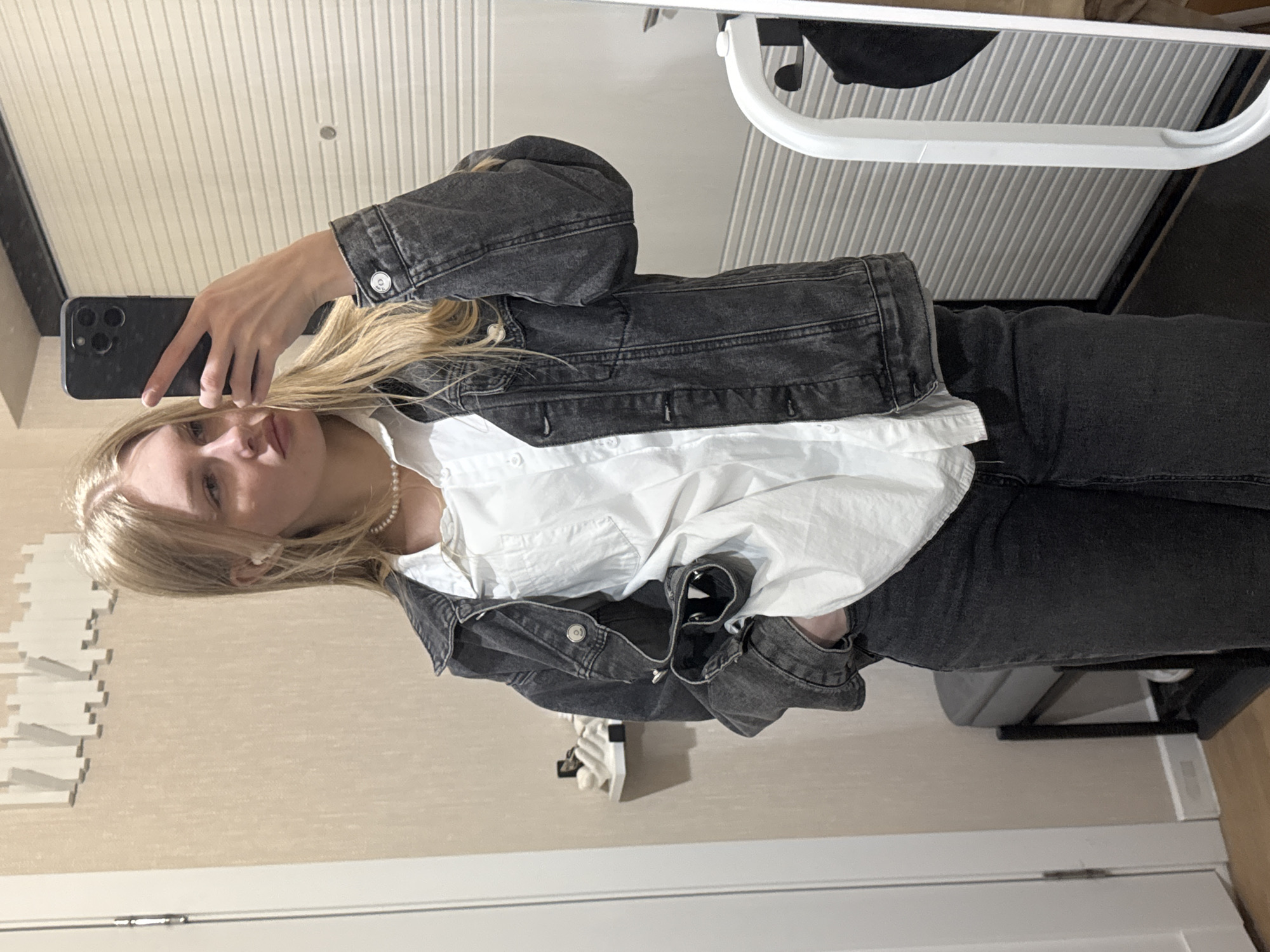 A young woman is captured in a mirror selfie, with the image rotated 90 degrees counter-clockwise, depicting her and the room sideways. She has blonde hair and a neutral expression, looking towards the camera (her smartphone). She holds a black smartphone, likely an iPhone, in her right hand to take the photo.
She is wearing a casual outfit that includes a white collared shirt, a dark gray or black denim jacket, and matching dark gray or black denim jeans. A white pearl necklace adorns her neck.
The indoor setting in Perm, Russia, features light beige walls and a white slatted ceiling. On the left wall, there is a decorative white element, possibly a series of small shelves or textured wall art. In the background on the right, elements of furniture or a clothes rack are visible, including a white curved frame and a striped surface. A white door frame is also seen at the bottom of the image. There is no visible text.