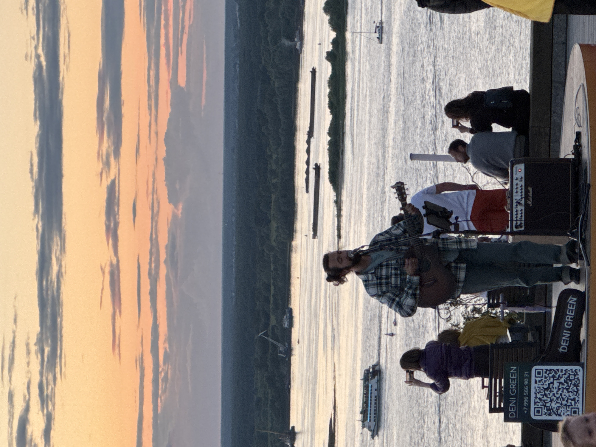 The image depicts an outdoor performance at sunset, likely in Nizhny Novgorod, Russia, given the presence of the iconic Nizhny Novgorod Kremlin visible in the distance. A man with his hair tied back is playing an acoustic guitar and singing into a microphone on a stage. He is wearing a plaid shirt and jeans. Behind him, a river flows with several boats, including a large passenger ferry, and barges. The sky is filled with hues of orange, pink, and gray clouds, indicating a sunset. Spectators are visible in the foreground and background, some holding up phones to capture the moment. A small amplifier with the "Marshall" logo is placed on the stage. A QR code and contact information for "DENI GREEN" are displayed on a sign on the stage. The general atmosphere is one of a relaxed, public event enjoying the twilight hours by the river.