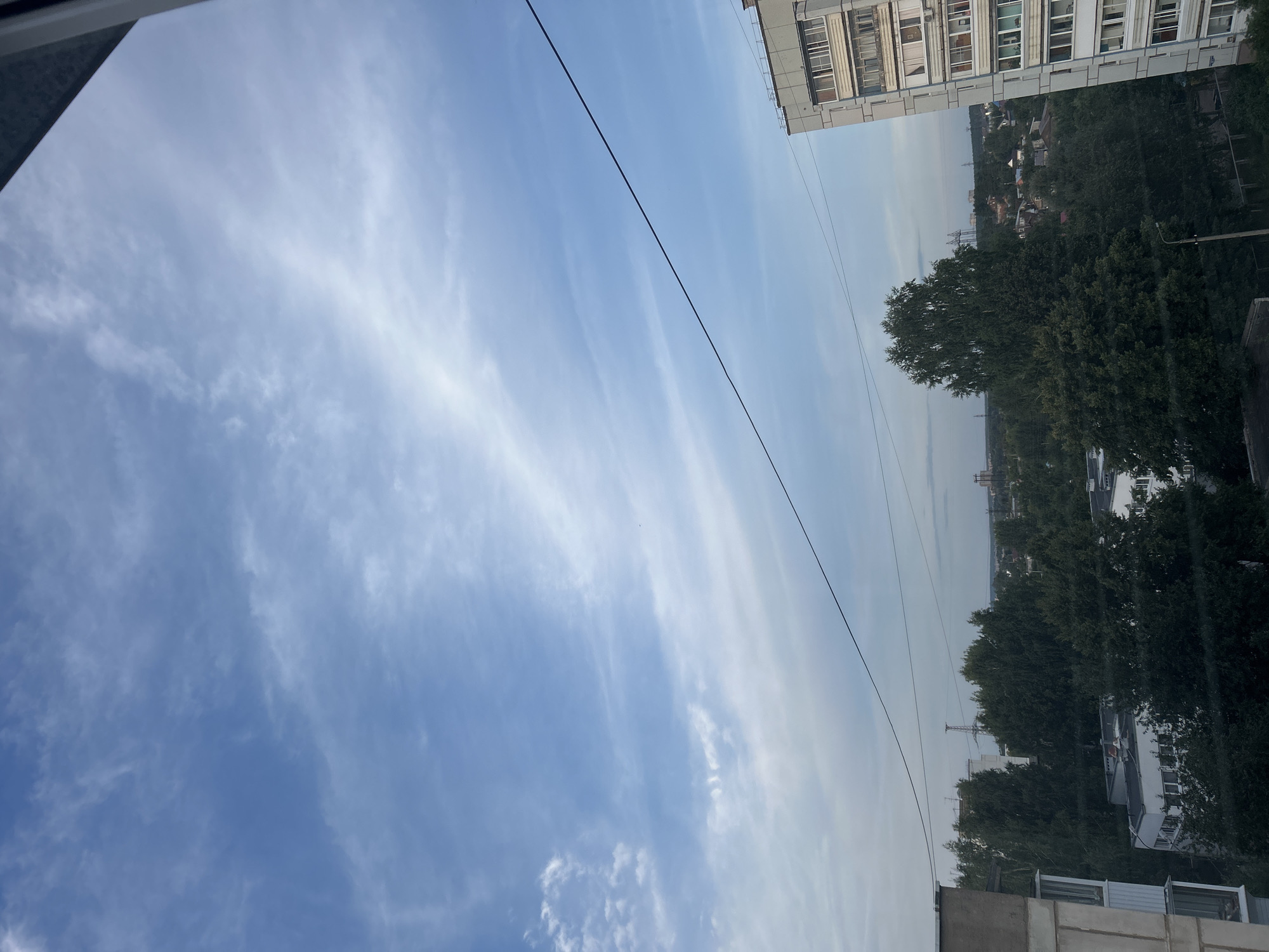 The image depicts a view from a high vantage point overlooking a city, likely Chaikovsky, Russia. The sky is a pale blue with wispy white clouds, suggesting a daytime scene. In the foreground, parts of two residential buildings are visible on the left and right edges. The building on the right is taller and appears to be a multi-story apartment block with balconies. A black electrical wire stretches diagonally across the frame from the upper right towards the center left. Below the sky, a dense canopy of dark green trees dominates the middle ground, interspersed with the roofs of lower buildings and houses. In the distance, more buildings and possibly industrial structures, including what appear to be power lines or pylons, are visible on the horizon. There are no people or animals clearly identifiable in the image.