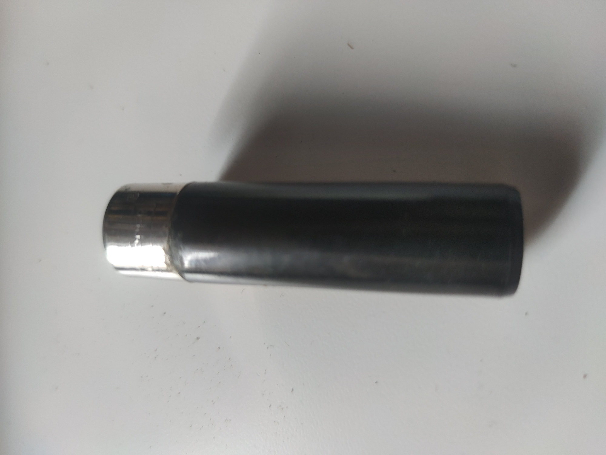 The image shows a cylindrical object with a black body and a silver cap, lying horizontally on a white surface. The object appears to be a flashlight or similar device. The environment is plain and lacks any distinct features that would indicate a specific location.