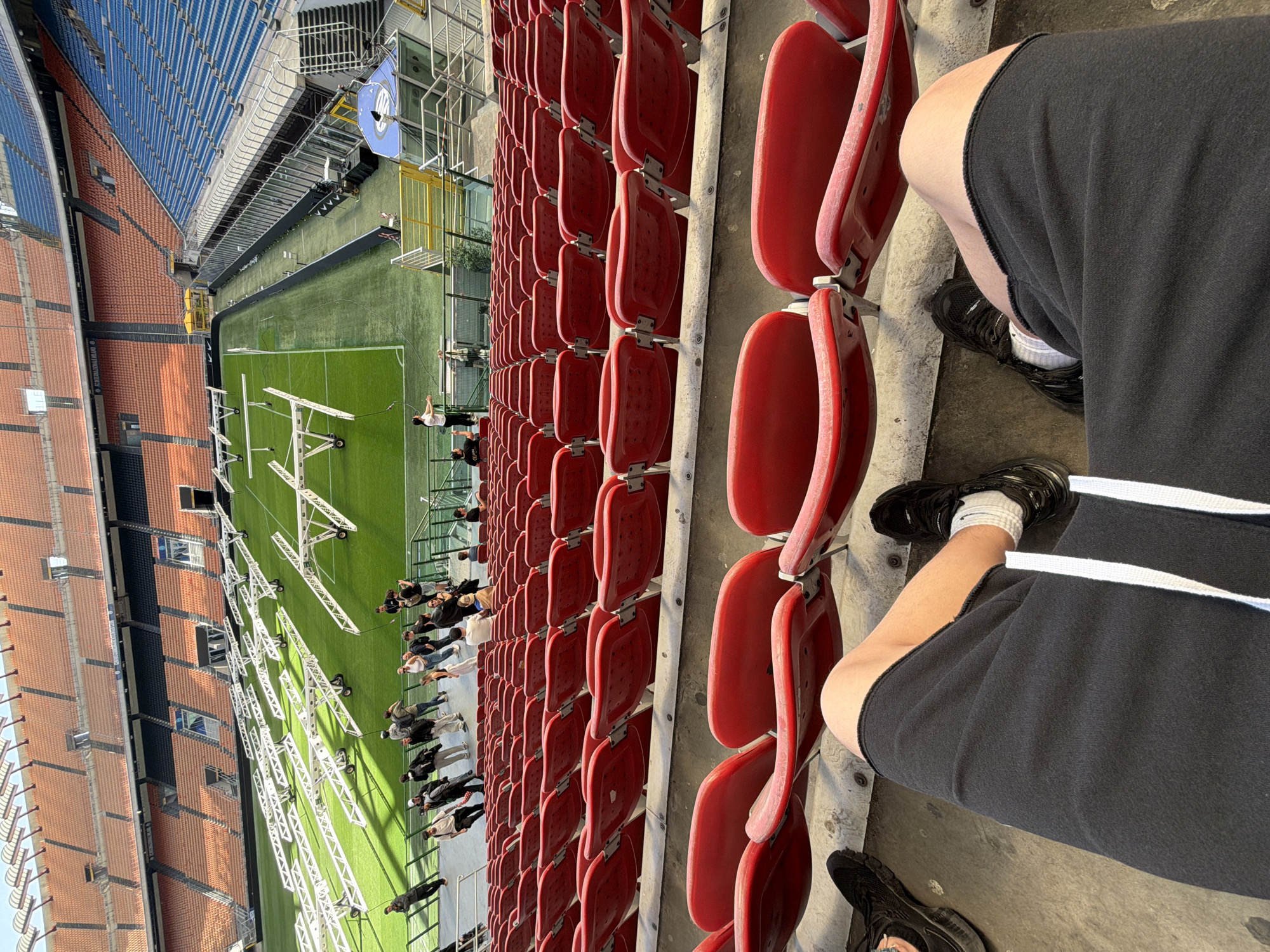 An elevated, slightly canted view of the San Siro stadium in Milan, Italy, showing a section of empty red seats and the adjacent pitch. The pitch is green, with a series of portable lighting rigs positioned across its surface. Several workers appear to be on the pitch, interacting with the equipment. In the foreground, a person is sitting in a stadium seat. They are wearing black shorts, a white sock, and black sneakers. The stadium architecture includes tiers of seating in blue and orange. The weather appears to be bright, suggesting a sunny day.