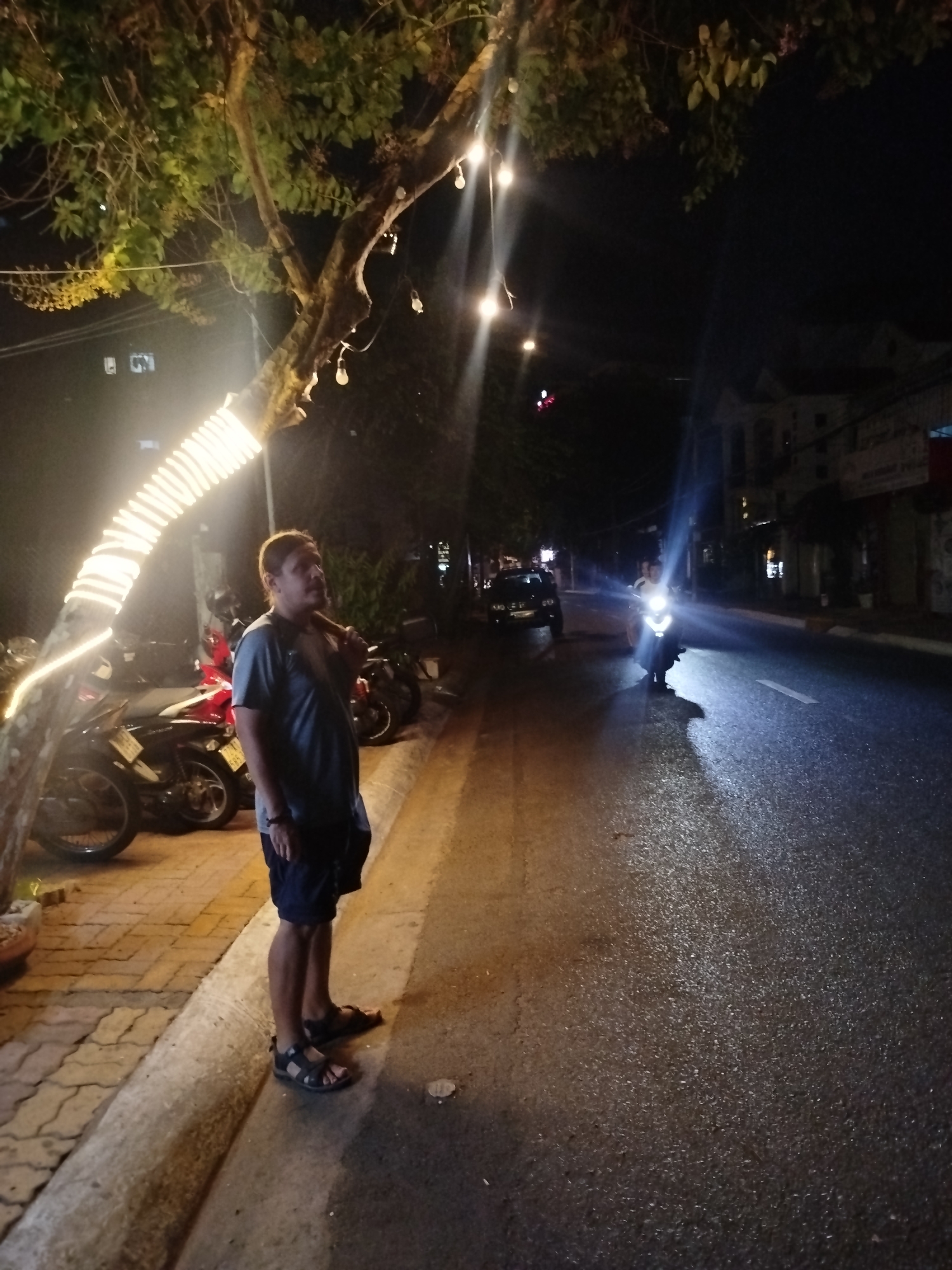 A man stands on a sidewalk next to a tree wrapped in lights. The scene is on a street in Ho Chi Minh City, Vietnam. Motorcycles are parked behind the man. Across the street, a person is riding a motorcycle towards the viewer. A car is parked further down the street. Buildings are visible in the background.