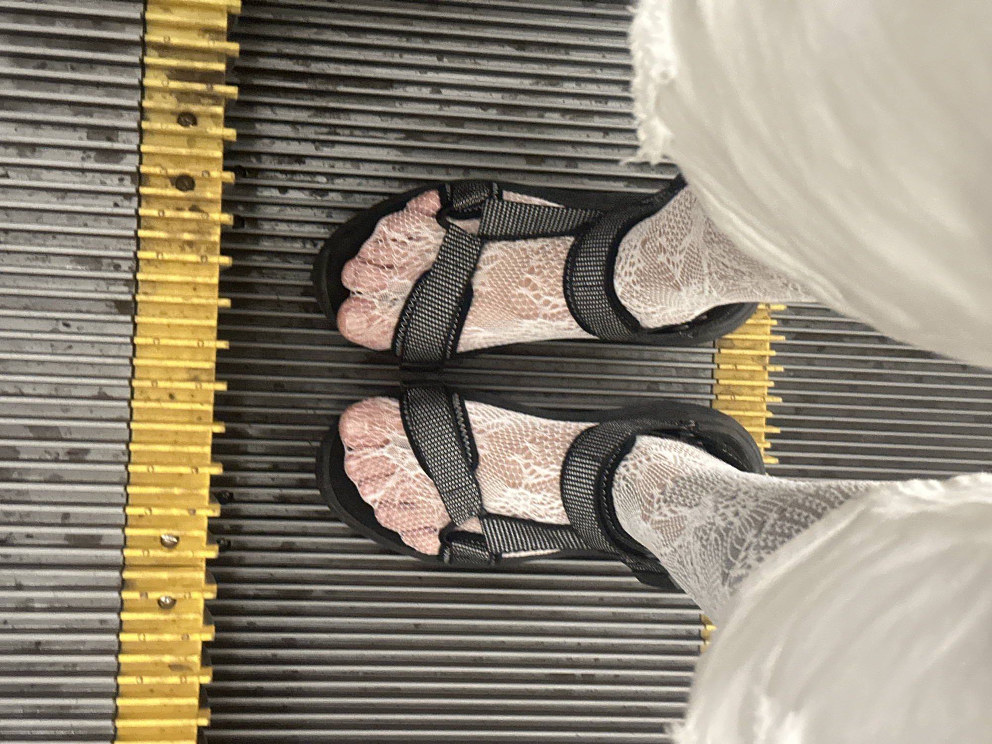 A top-down view shows two feet wearing black Teva-style sandals over white lace socks. The feet are positioned on an escalator. The escalator steps are ribbed, alternating between grey and yellow. The image is devoid of any identifying information that could place it in Moscow, Russia. The focus is solely on the footwear and the escalator.