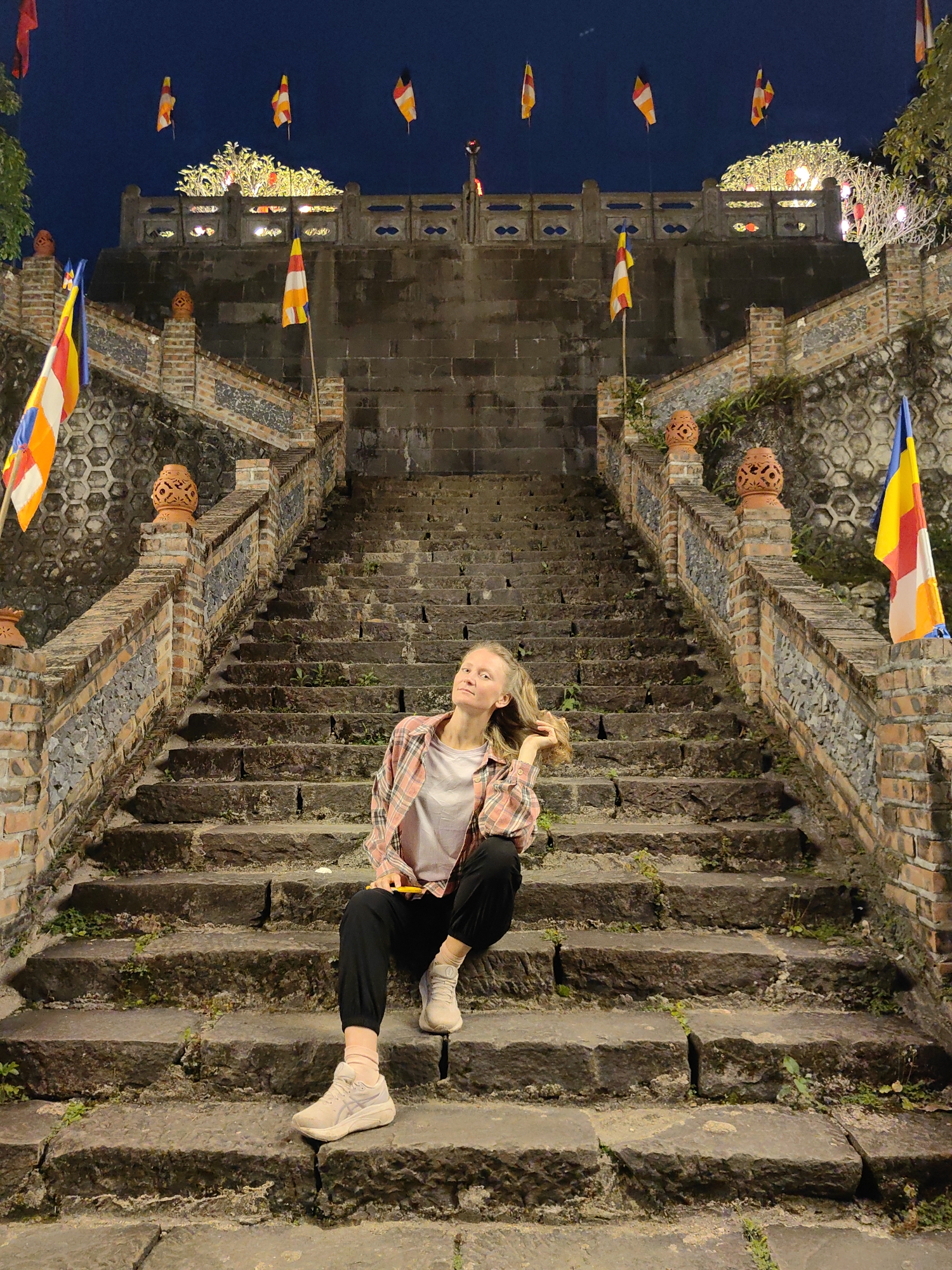 A woman is sitting on stone steps in what appears to be an outdoor historical or religious site in Xã Đàm Thủy, Vietnam. The steps ascend to a stone wall with decorative lights and flags on poles. The woman is wearing a plaid shirt, a plain t-shirt, dark pants, and sneakers. She is sitting with her legs extended and her left hand is touching her hair. The steps are weathered and have some greenery growing between the stones. Flags are placed along the walls lining the steps. The sky is dark, suggesting it is nighttime.