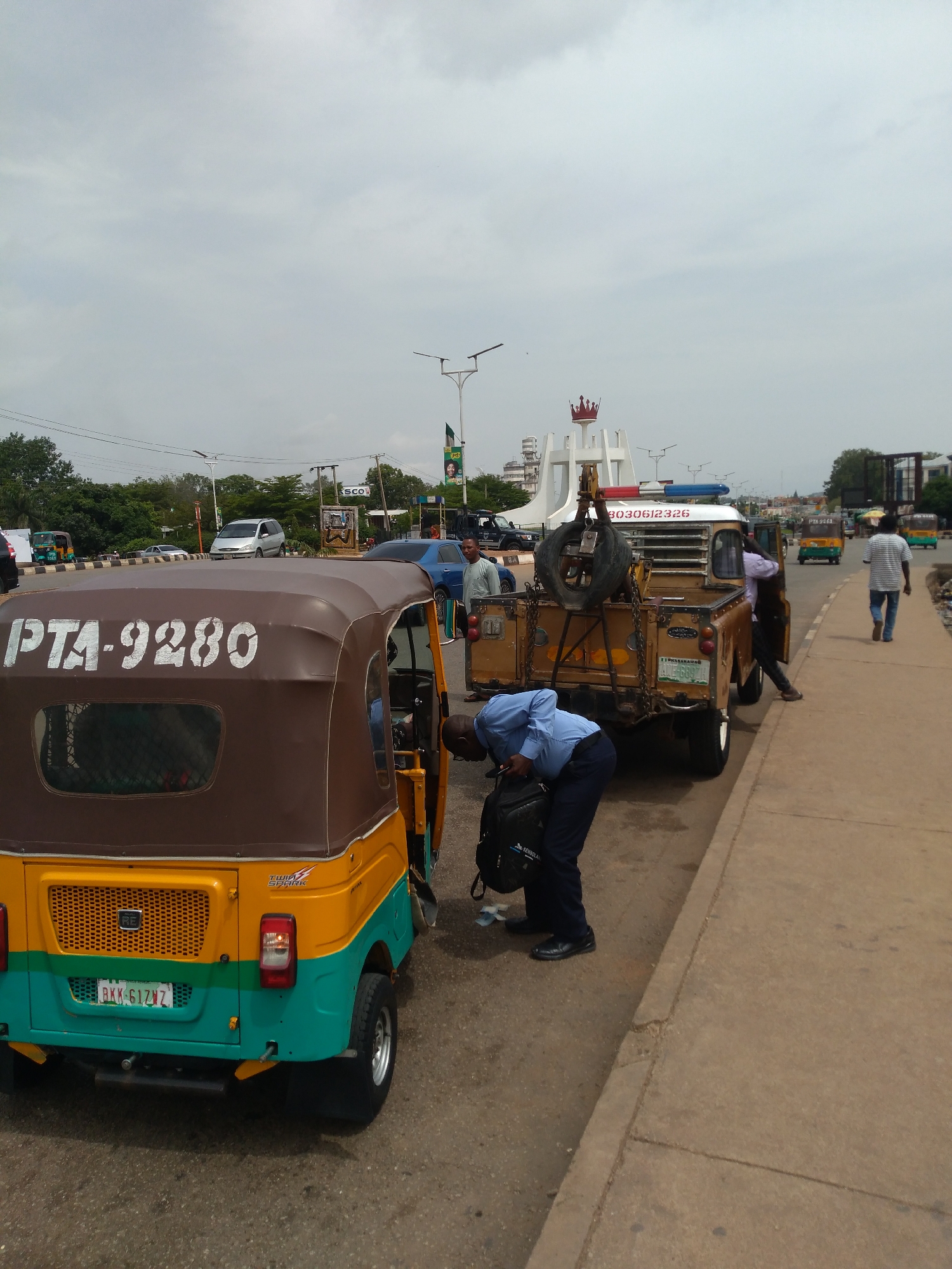 The image depicts a street scene in Jos, Nigeria. In the foreground, a yellow and teal auto-rickshaw with the registration plate "PTA-9280" is parked. A man in a blue uniform is bending down, placing a black backpack on the ground next to the auto-rickshaw. Behind the auto-rickshaw, a tan tow truck with a spare tire mounted on the back and a winch is positioned. A person in a light purple shirt is leaning into the tow truck. Further down the street, several other auto-rickshaws and a blue car are visible. In the background, there is a white monument topped with a red crown, streetlights, and trees, indicating an urban setting. The sky is overcast.