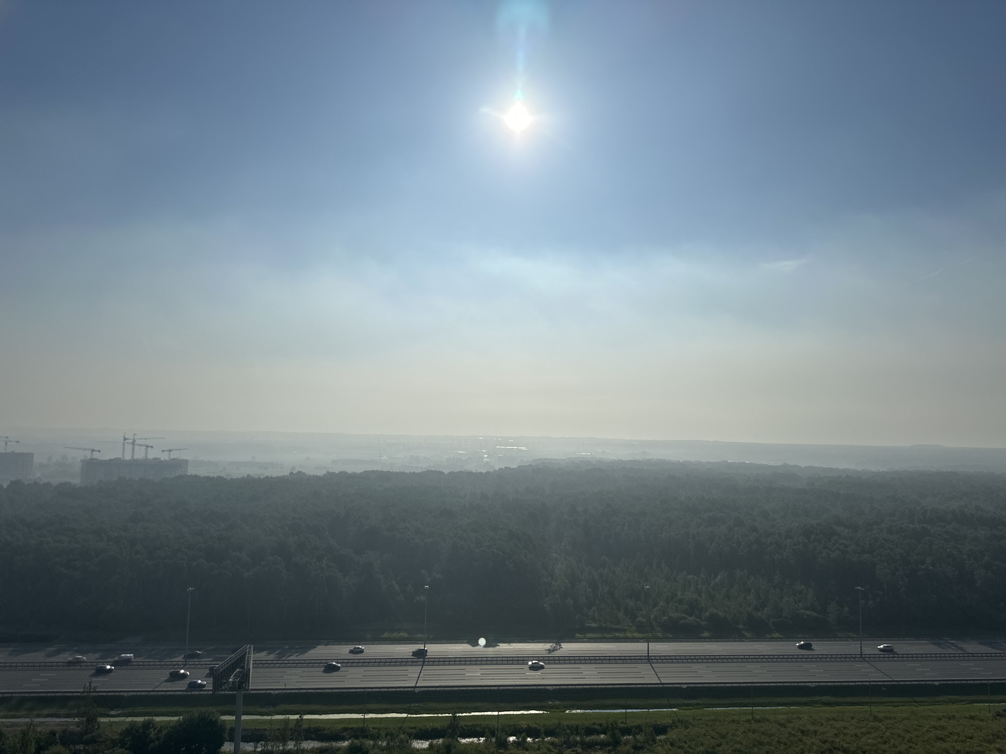 This is a wide shot aerial photograph of a landscape that includes a highway, a dense forest, and a city under construction in the distance, bathed in the bright glare of the sun at the top of the frame. The scene is set in the city of Nikolskoye, Russia.
The foreground of the image features a multi-lane highway with several cars driving on it. The highway is bordered by green grass and some trees. Behind the highway, a vast expanse of dark green forest stretches across the middle ground, with the trees appearing dense and uniform. In the far distance, a hazy cityscape is visible, with construction cranes and buildings indicating ongoing development. The sky is a pale blue, with a bright, star-like sun emitting a lens flare at the center. The overall atmosphere is hazy, suggesting either early morning mist or light pollution.
