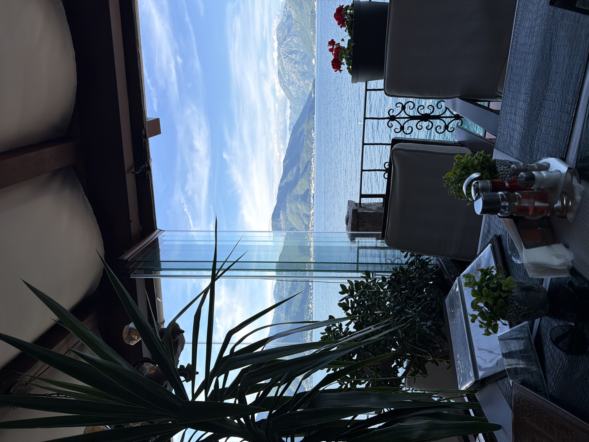 The image depicts a partially obscured view from a restaurant or balcony in Krašići, Montenegro, facing the Bay of Kotor. A mountain range dominates the distant backdrop. The foreground features interior elements including a glass partition, seating, plants, and table settings with condiments. Sunlight illuminates parts of the scene.