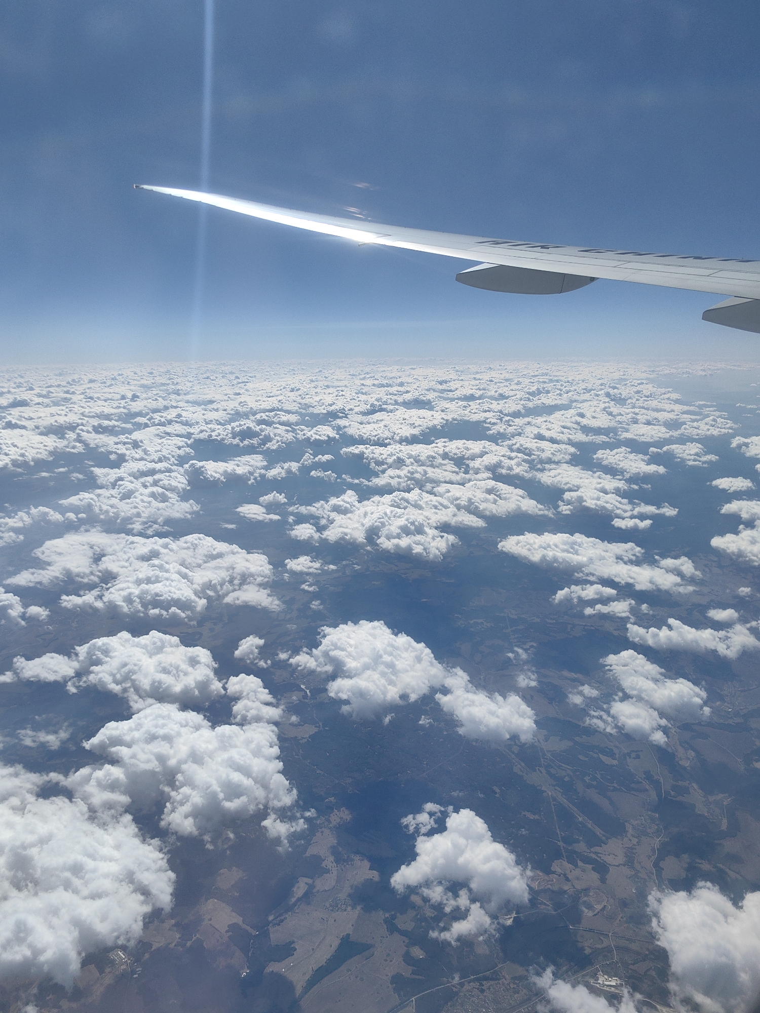 The image shows a view from an airplane window, looking down at a landscape covered partially by clouds. The airplane wing is visible in the upper right and upper center of the frame. Below, there is a layer of puffy white clouds, with patches of land visible in between the clouds. The land appears to be a mix of fields and possibly wooded areas. It is not possible to definitively identify the city of Novotroitsk, Russia, based solely on this image.