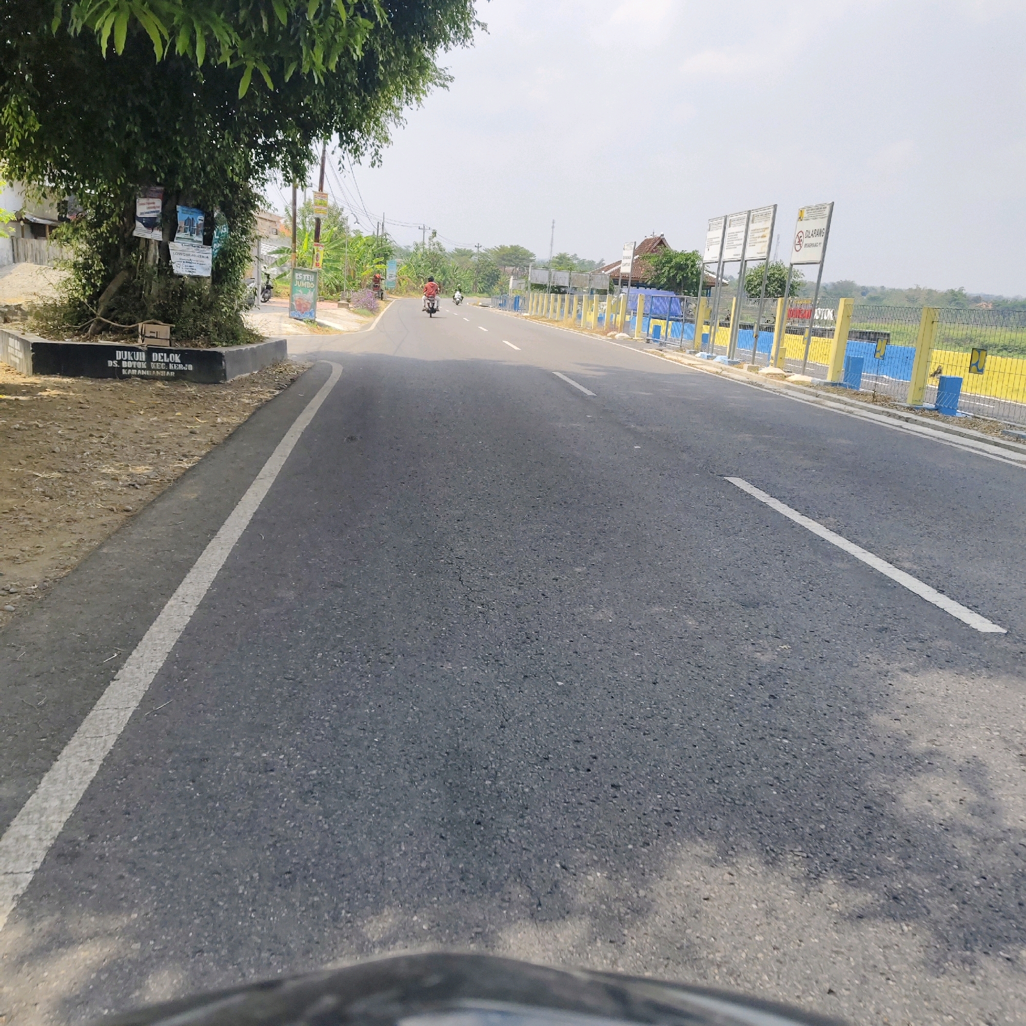 The image depicts a road in the city of Botok, Indonesia. The road is paved and has a white line marking the edge of the lane. On the left side of the road, there is a large tree with several posters attached to its trunk. A black concrete structure with white lettering is visible at the base of the tree, indicating "DUKUN DELOK, DS. BOTOK, KEC. KERJO, KARANGBAR". The background shows a fence with blue and yellow posts, and beyond that, a field of vegetation. Several signs are mounted on poles along the side of the road. In the distance, there are a few buildings and more trees, with a hazy sky overhead. Two people on motorcycles are traveling down the road, one in the foreground and one further ahead. The scene appears to be an outdoor environment with clear daylight.