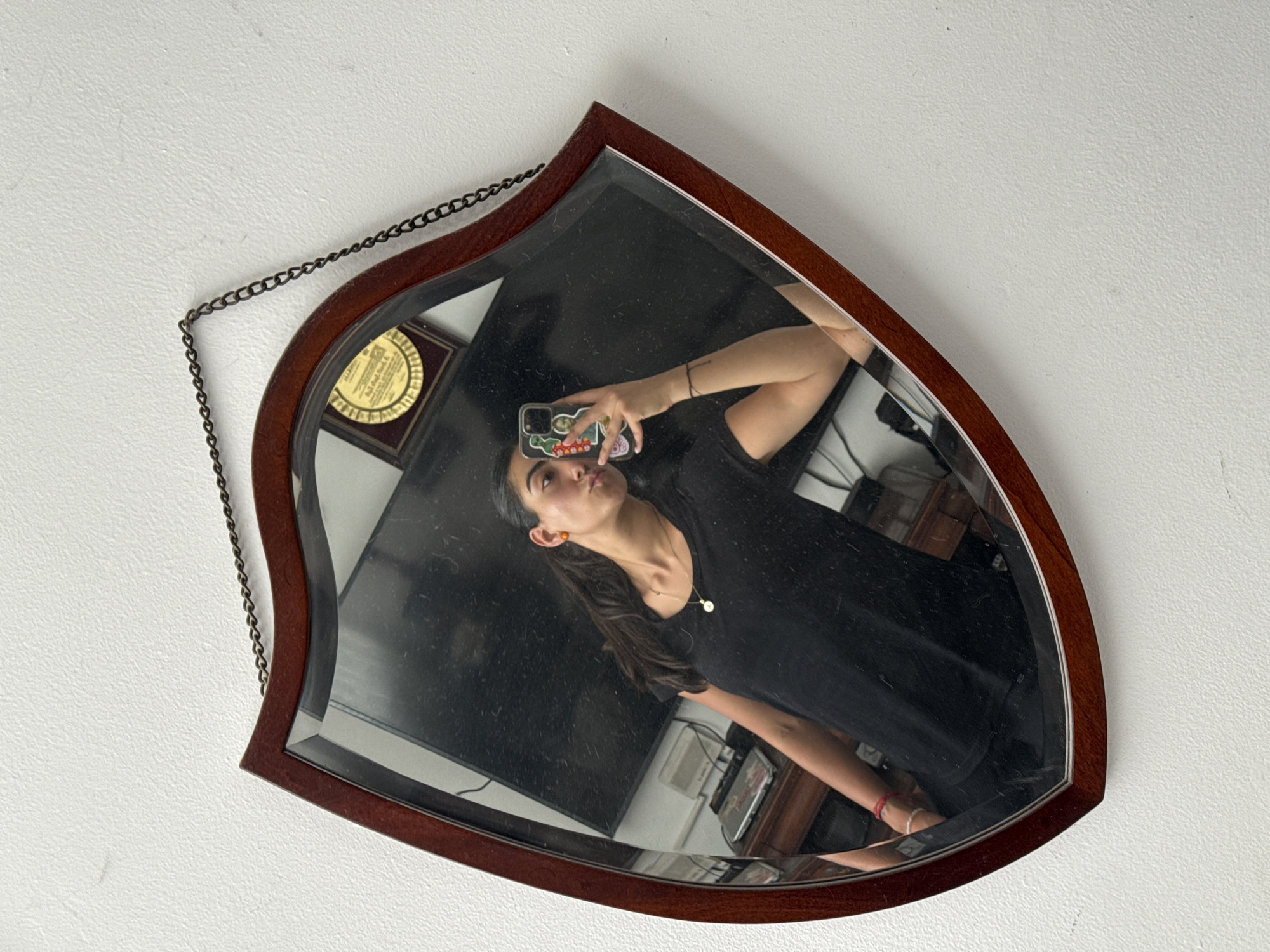 A person is reflected in a shield-shaped, wood-framed mirror hanging on a white wall. The person is holding a phone with stickers on the back, taking a selfie. The reflection shows a dark-haired person wearing a dark-colored shirt and a gold necklace. Behind the person in the reflection is a TV, a shelf, and various other objects, suggesting an interior setting. A gold-colored plaque with a dark border is also visible in the reflection, mounted behind the person. A chain hangs from the mirror's top left corner. There is no indication that this is Puebla, Mexico.