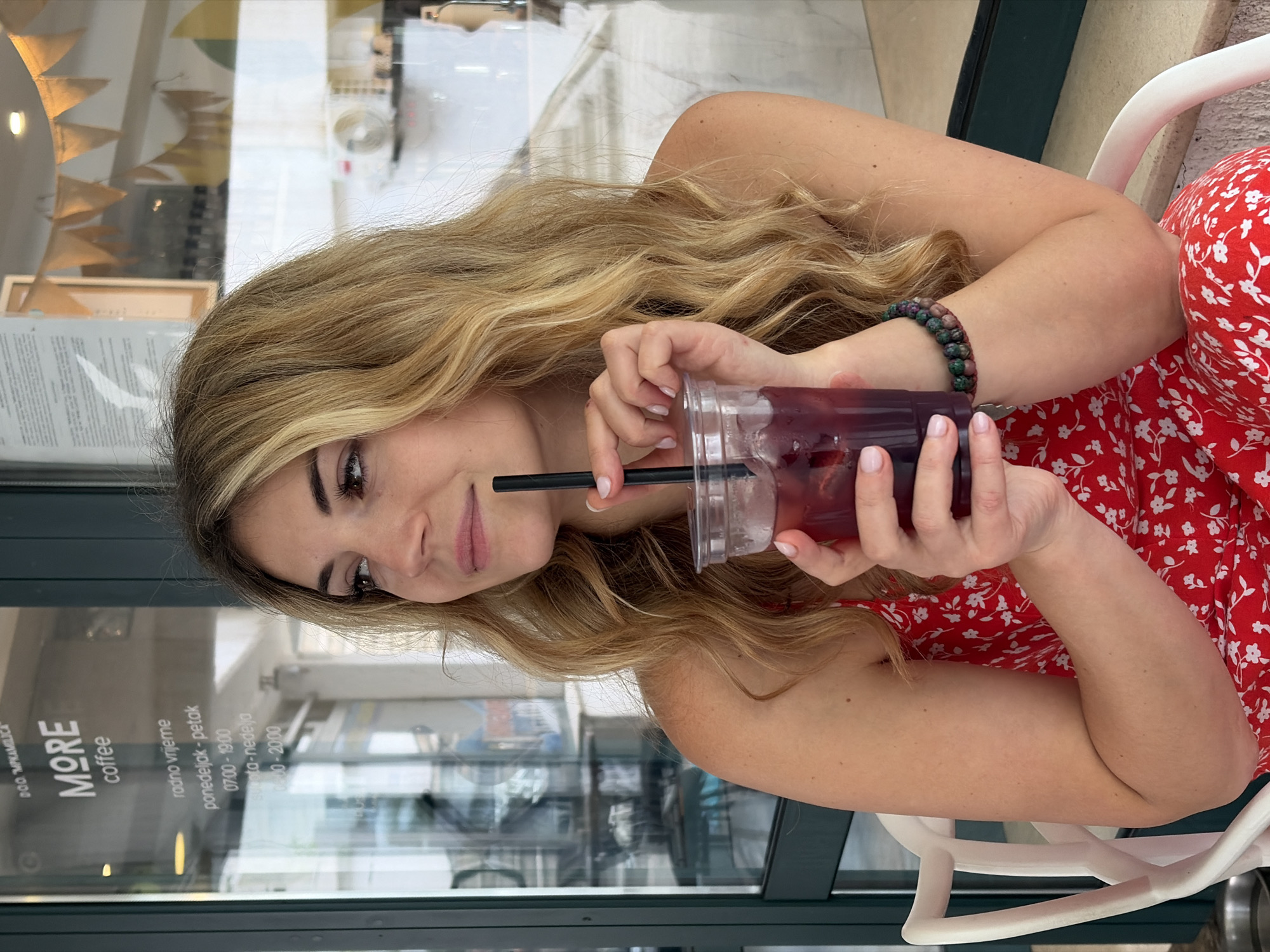 The image shows a woman with long, blonde wavy hair and a red and white floral dress sitting in a cafe. She is holding a plastic cup filled with a dark beverage and ice, using a black straw. The cafe appears to be named "MORE coffee", as indicated on the glass window behind her. The opening hours of the cafe are also displayed, suggesting it's in Budva, Montenegro. The woman wears a beaded bracelet on her left wrist. She looks casually towards the camera. The overall atmosphere seems relaxed, possibly during a casual outing in the city.