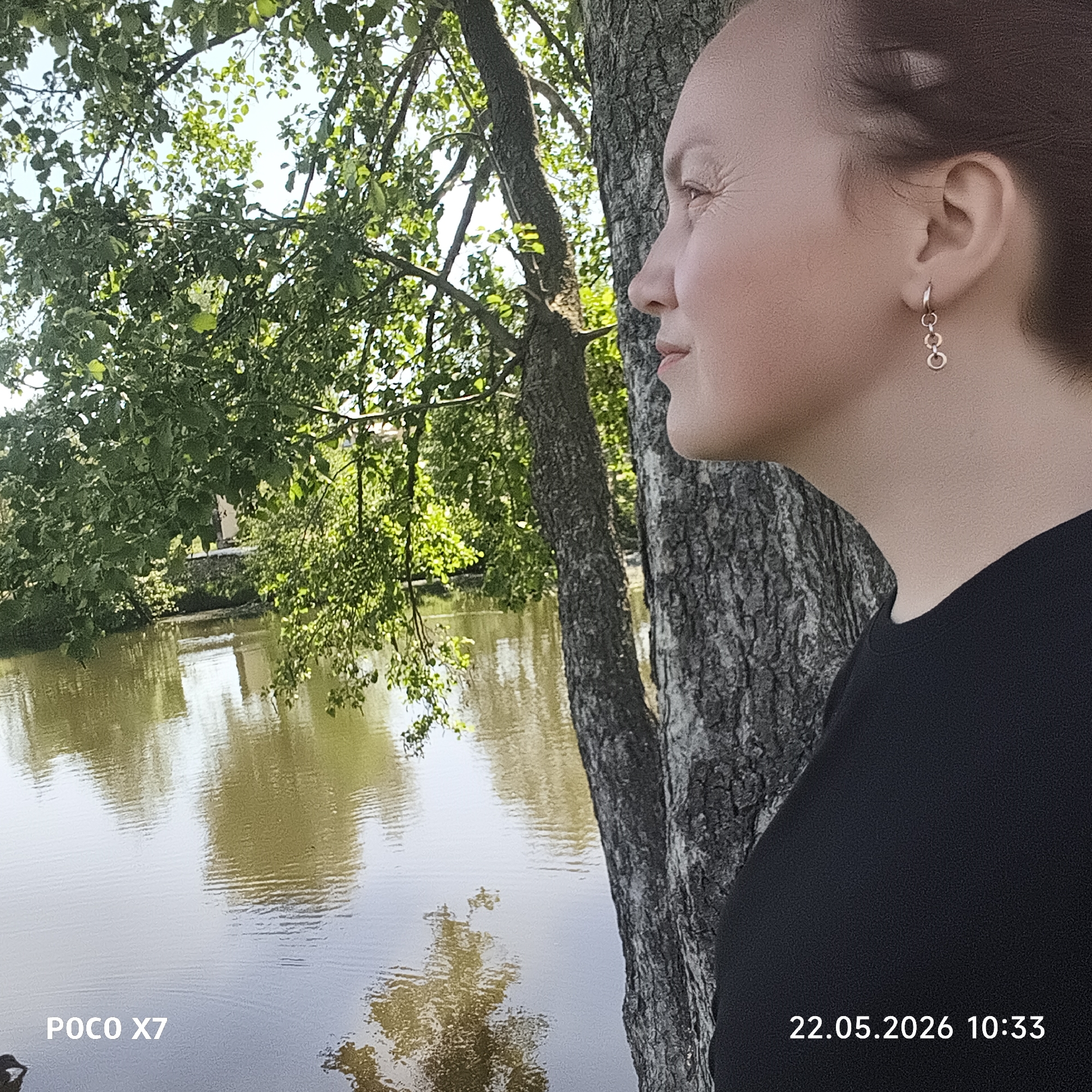 A woman stands near a tree next to a body of water on May 22, 2026 at 10:33, as recorded by a POCO X7 camera. The location appears to be in the city of Chaikovsky, Russia, as indicated by the date format. The woman has reddish-brown hair and is wearing earrings. She is looking toward the water, which reflects the trees and sky. A bridge can be seen in the distance.