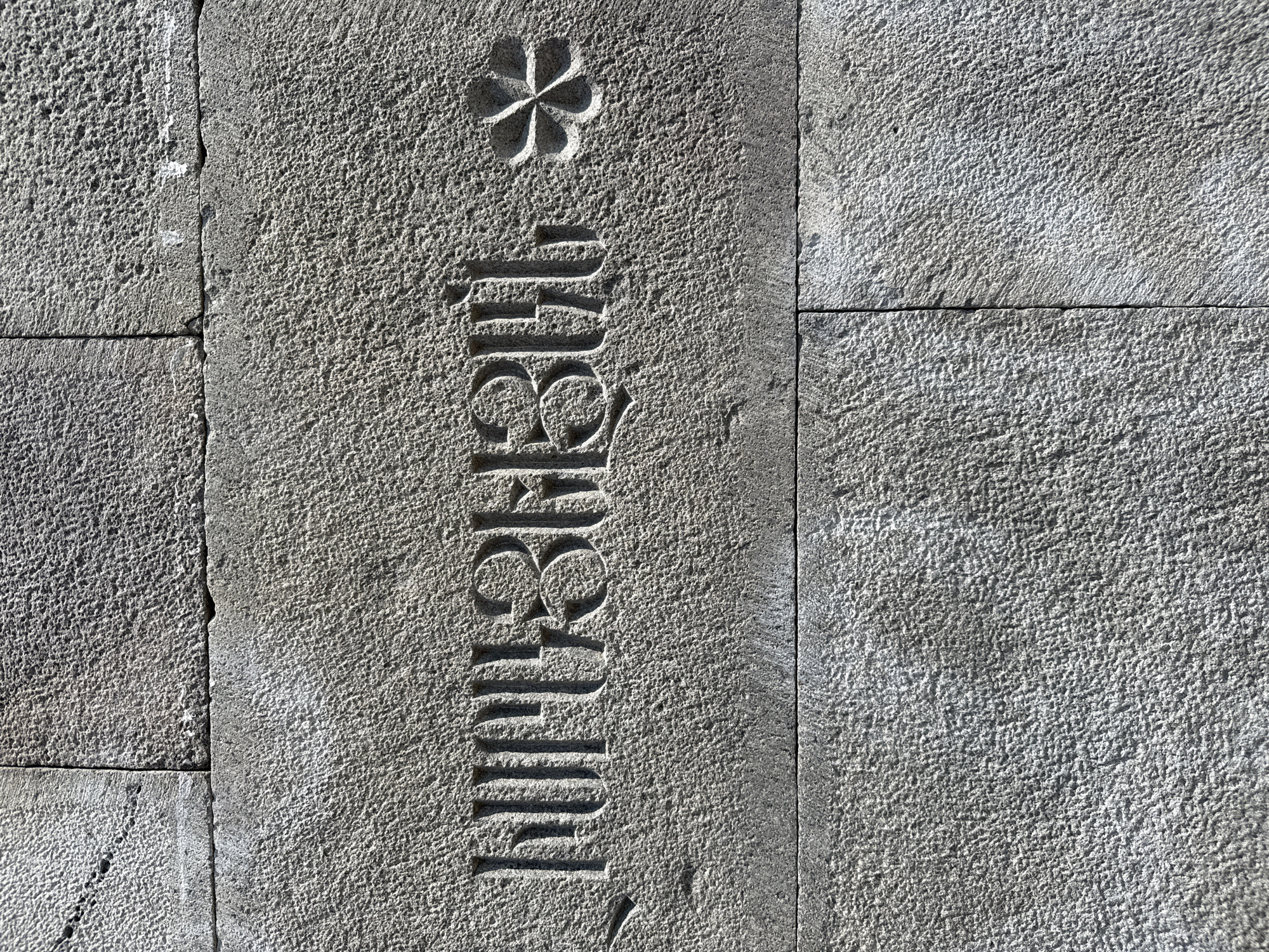 The image shows a section of a stone wall with carved Armenian script. The text is carved into a light grey, rough-textured stone block, which is part of a larger wall constructed from similarly textured blocks. To the right of the text, there is a carved floral motif with four petals. The wall is located in the city of Voghjaberd, Armenia. The lighting suggests it is daytime, with shadows indicating the direction of the sun. No people or animals are visible in the image.