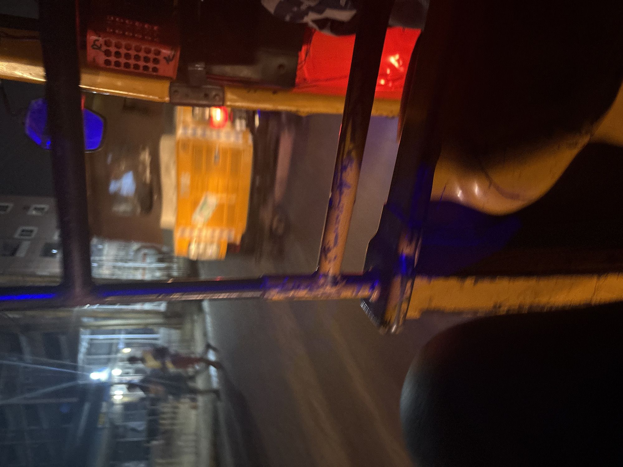 The image captures a nighttime view from within an auto-rickshaw, likely in Nkpor, Nigeria. In the foreground, a dark, curved surface, possibly a seat, is visible. A yellow metal bar runs across the middle ground, illuminated by reflections of blue and red lights. Beyond the bar, the interior of another vehicle, possibly a truck or another auto-rickshaw, is seen with a bright yellow cargo. A red taillight is visible on this vehicle. In the background, a street scene unfolds with blurred figures of people walking, illuminated by streetlights. Buildings line the street. The overall impression is of a busy urban environment at night, possibly in motion given the perspective.