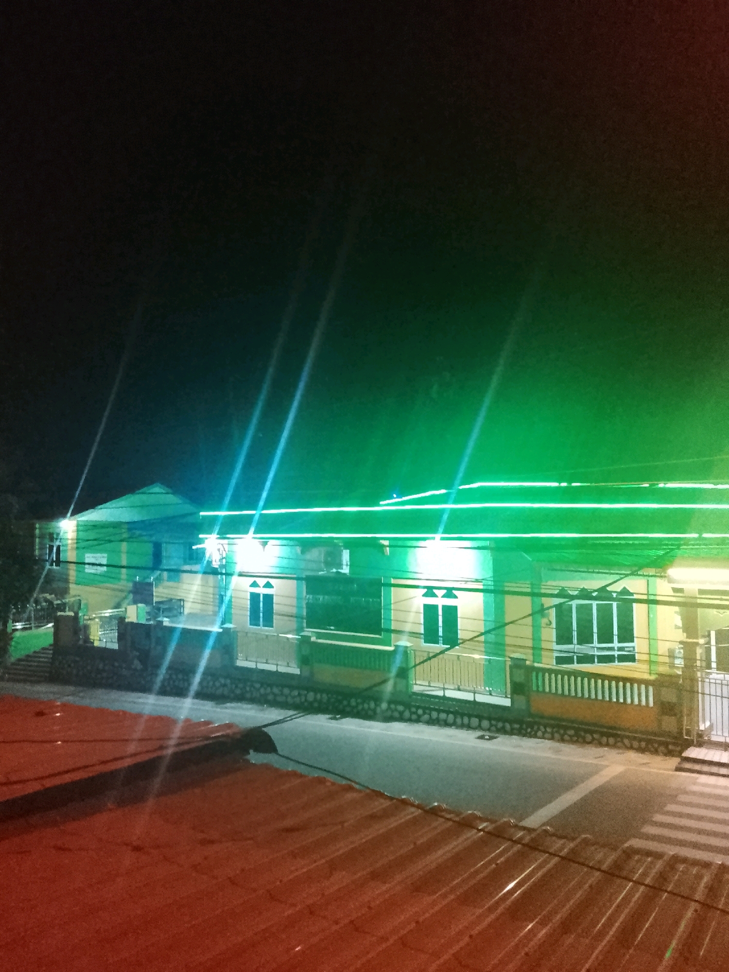 The image shows a two-story building illuminated by bright green neon lights at night. The building's facade is painted yellow and green, with white window frames. A paved road runs in front of the building, featuring a pedestrian crossing. In the foreground, a red corrugated metal roof is visible, casting shadows from the building. The scene appears to be in a residential or commercial area within the city of Tanjung Balai Karimun, Indonesia. The overall atmosphere is one of nighttime illumination and urban scenery.