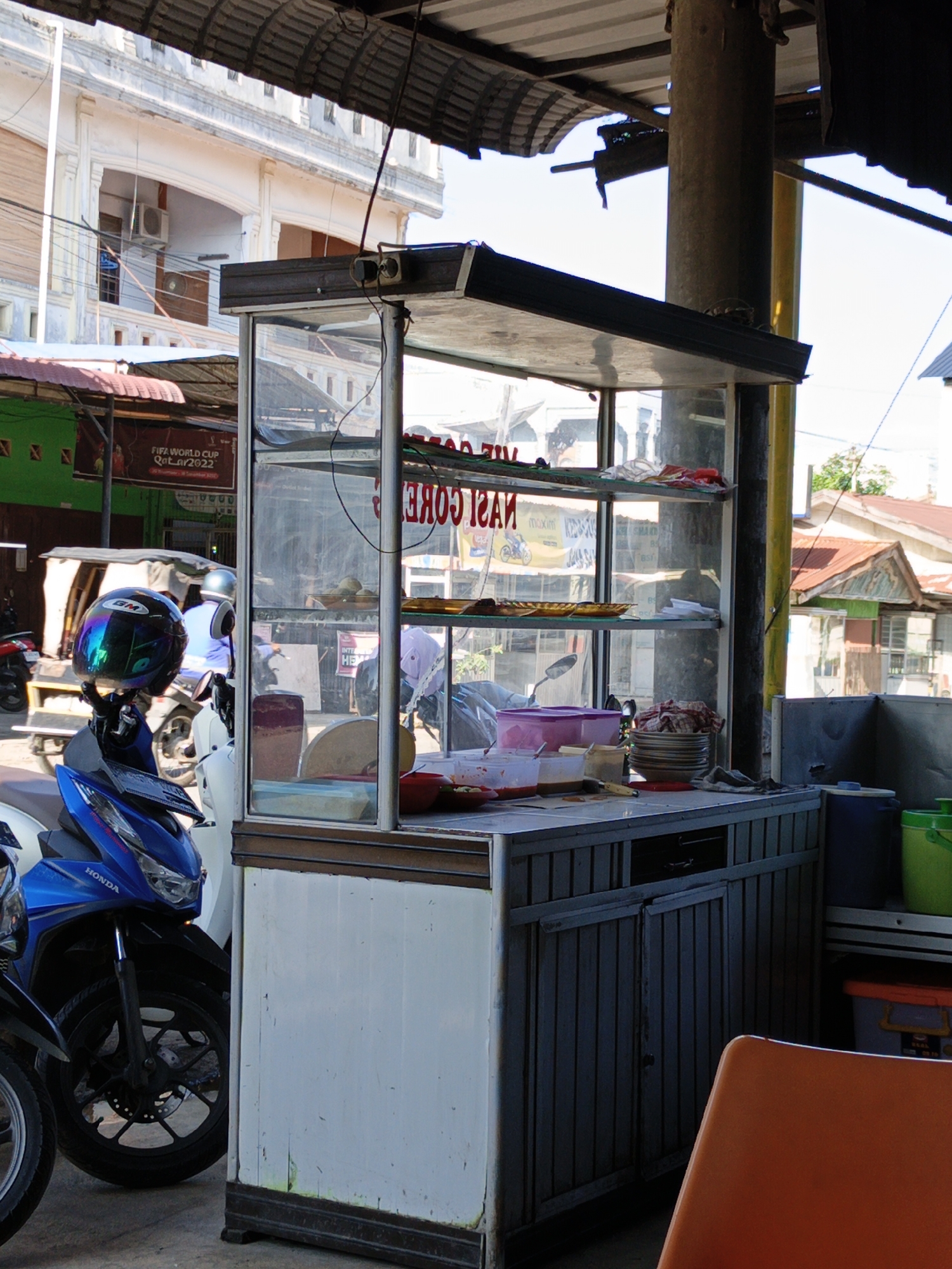 The image shows a street food stall in Lhokseumawe, Indonesia. A glass-enclosed display case sits on a cabinet with a counter. The shelves inside hold food, plates, and utensils. The stall is positioned under an awning, supported by a large concrete pillar. In the foreground, a blue Honda motorcycle is parked near the stall. In the background, buildings are visible across the street, including a green building with a FIFA World Cup Qatar 2022 banner and a white building with architectural details. An orange chair is partially visible in the lower right corner.