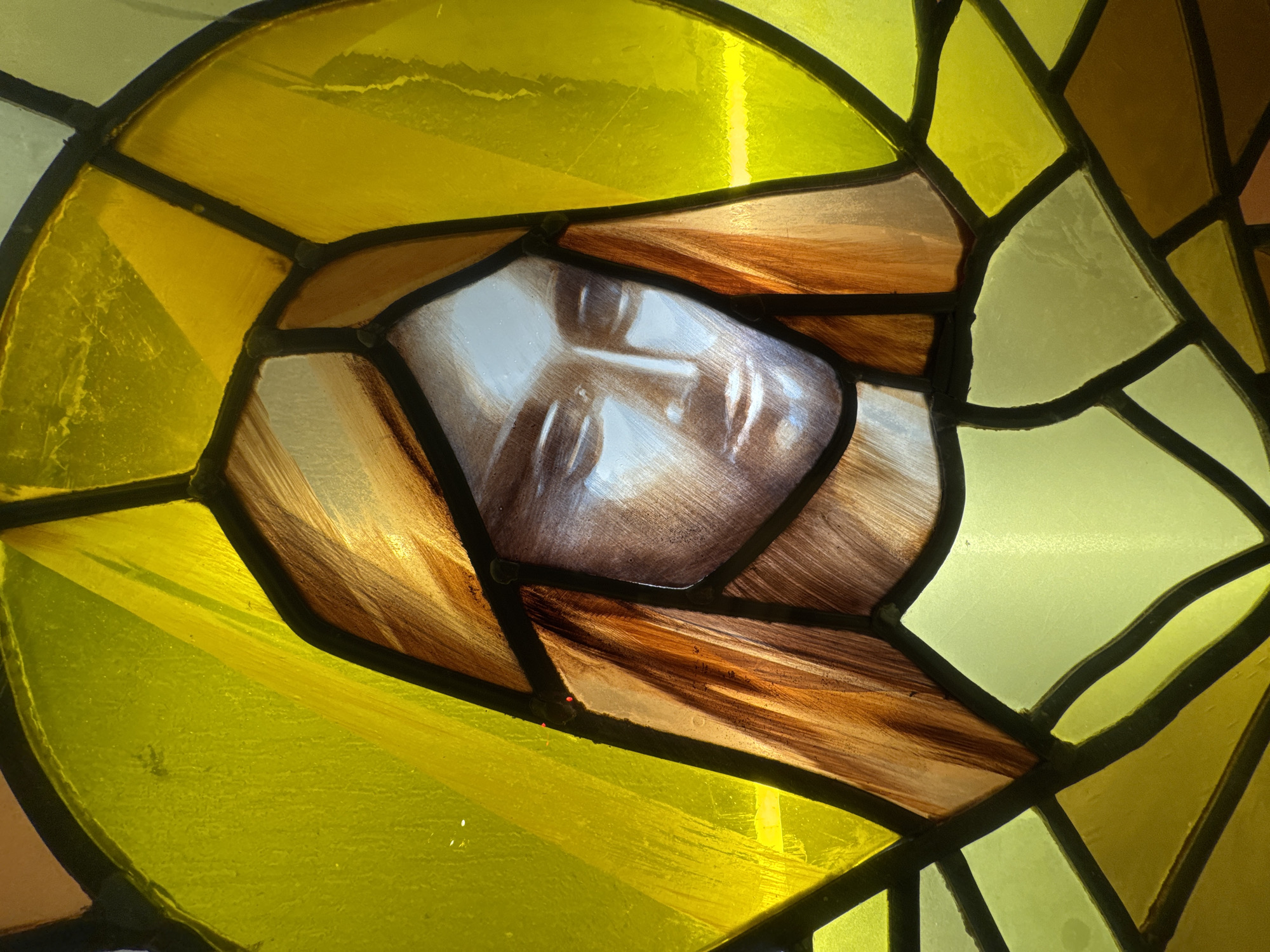 A section of stained glass artwork features a face in the center, surrounded by pieces of yellow, brown, and clear glass. The glass is separated by black lines. No information about the location in Paris is available from the image.