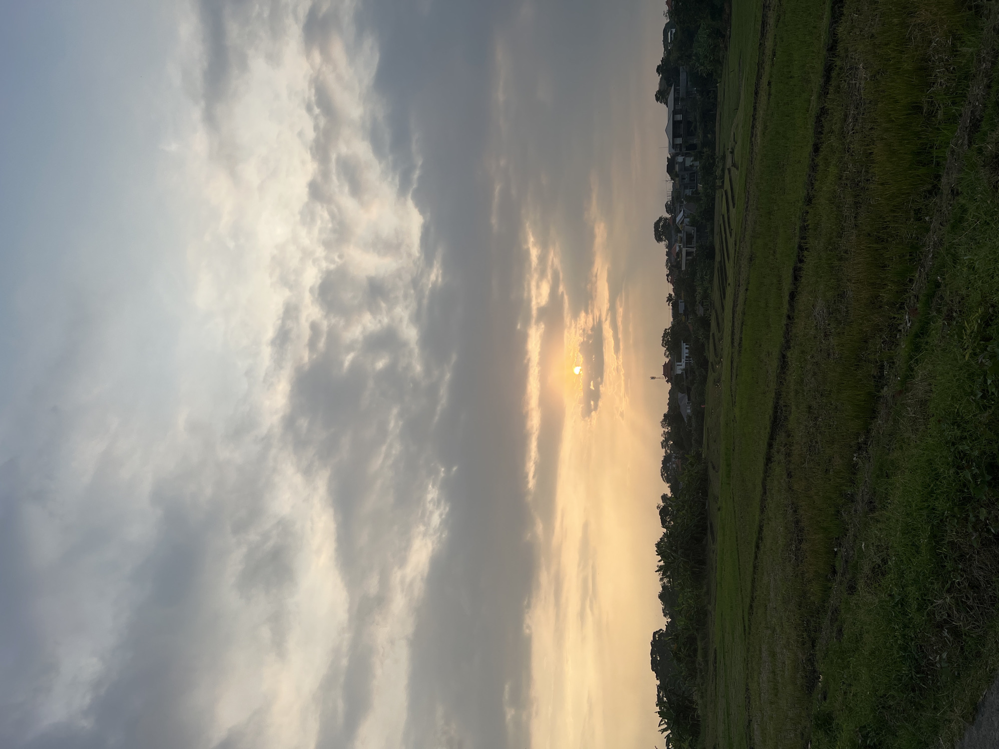 The image shows a landscape at dusk or dawn in North Kuta, Indonesia, featuring a field of green vegetation in the foreground. The sky is filled with clouds, some grey and others illuminated by the sun, which is partially obscured by the clouds. Buildings are visible in the distance behind the vegetation.