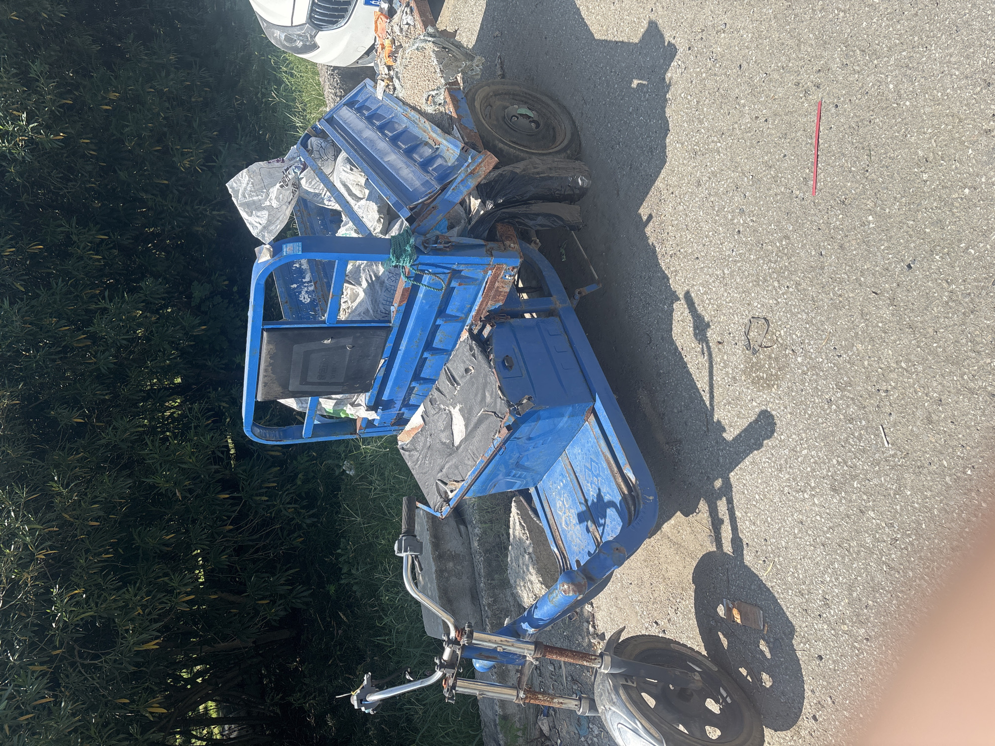 An abandoned, three-wheeled electric scooter with a blue metal chassis and a black seat is parked on an asphalt road. The scooter is loaded with white sacks and what appears to be debris. The front of the scooter has a single wheel and handlebars, while the rear has two wheels and a cargo bed. The scooter is weathered and shows signs of rust. In the background, there is a dense green foliage and a portion of a white car. The scene appears to be located outdoors, possibly in a semi-urban or industrial area.