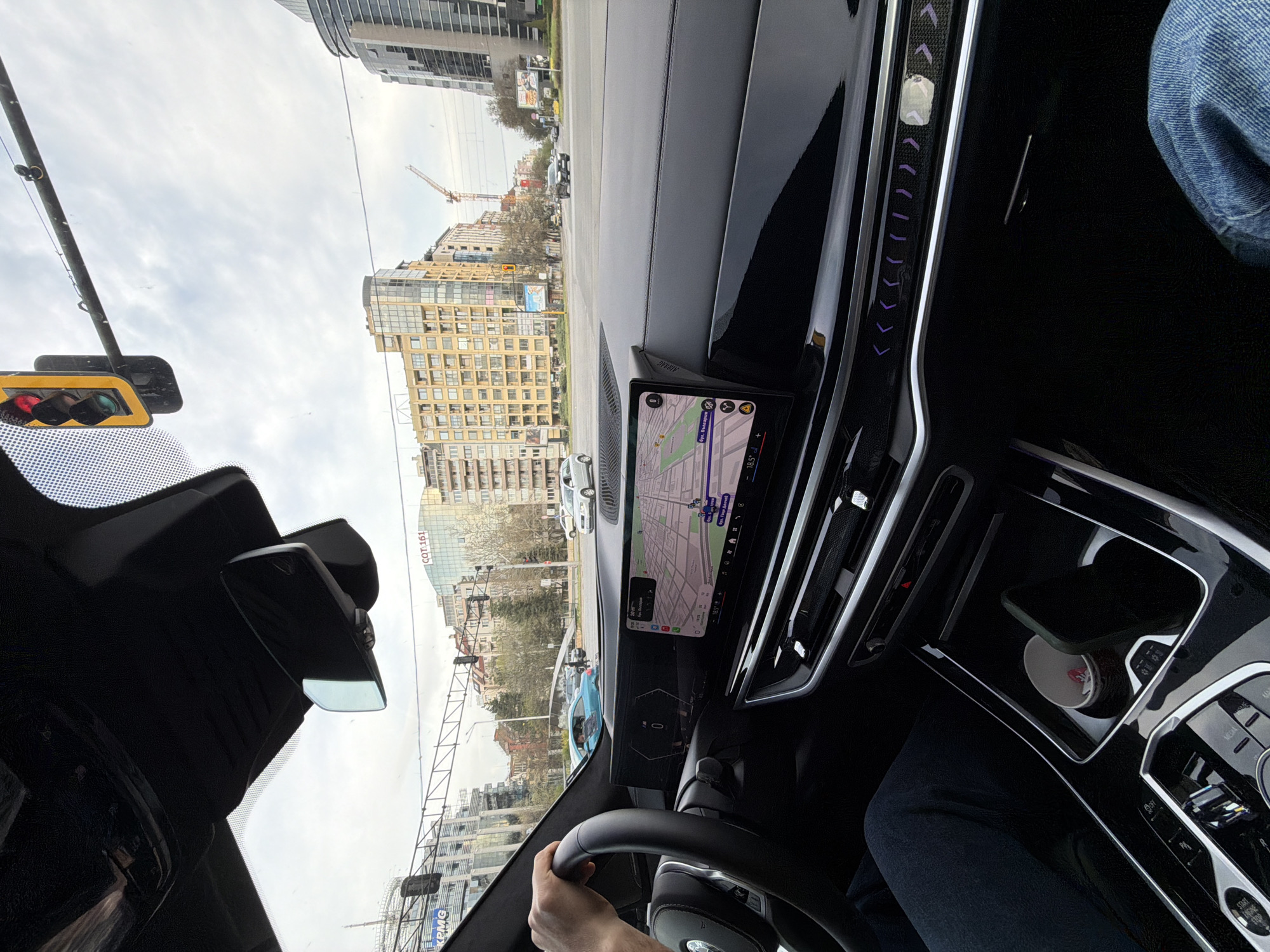 The image captures the interior of a modern car, likely a BMW given its distinctive curved display and cabin design, from the driver's perspective, looking out onto a city street in Sofia, Bulgaria. A person's hand is visible on the steering wheel, and their leg, dressed in jeans, is seen in the lower right.
The car is stationary, with a traffic light showing red and green signals visible through the windshield, suggesting it is waiting at an intersection. The car's central display shows a navigation map with Cyrillic street names "бул. Гоце Делчев" and "бул. България," which are major boulevards in Sofia. The display also shows an ambient temperature of 18.5°C. The digital instrument cluster indicates "0" for speed. On the passenger side, a mobile phone rests in a cupholder next to a small white cup. The dashboard features sleek, illuminated accents with arrow patterns and an "AIRBAG" label.
Outside the car, multi-story buildings, some appearing commercial or residential, line the street under an overcast sky. A construction crane is visible in the distance, indicating ongoing development. Signs for "KPMG" and "COT 161" are identifiable on some of the buildings. Other vehicles are visible on the road. The time of day appears to be daytime, with soft, diffused light from the cloudy conditions.